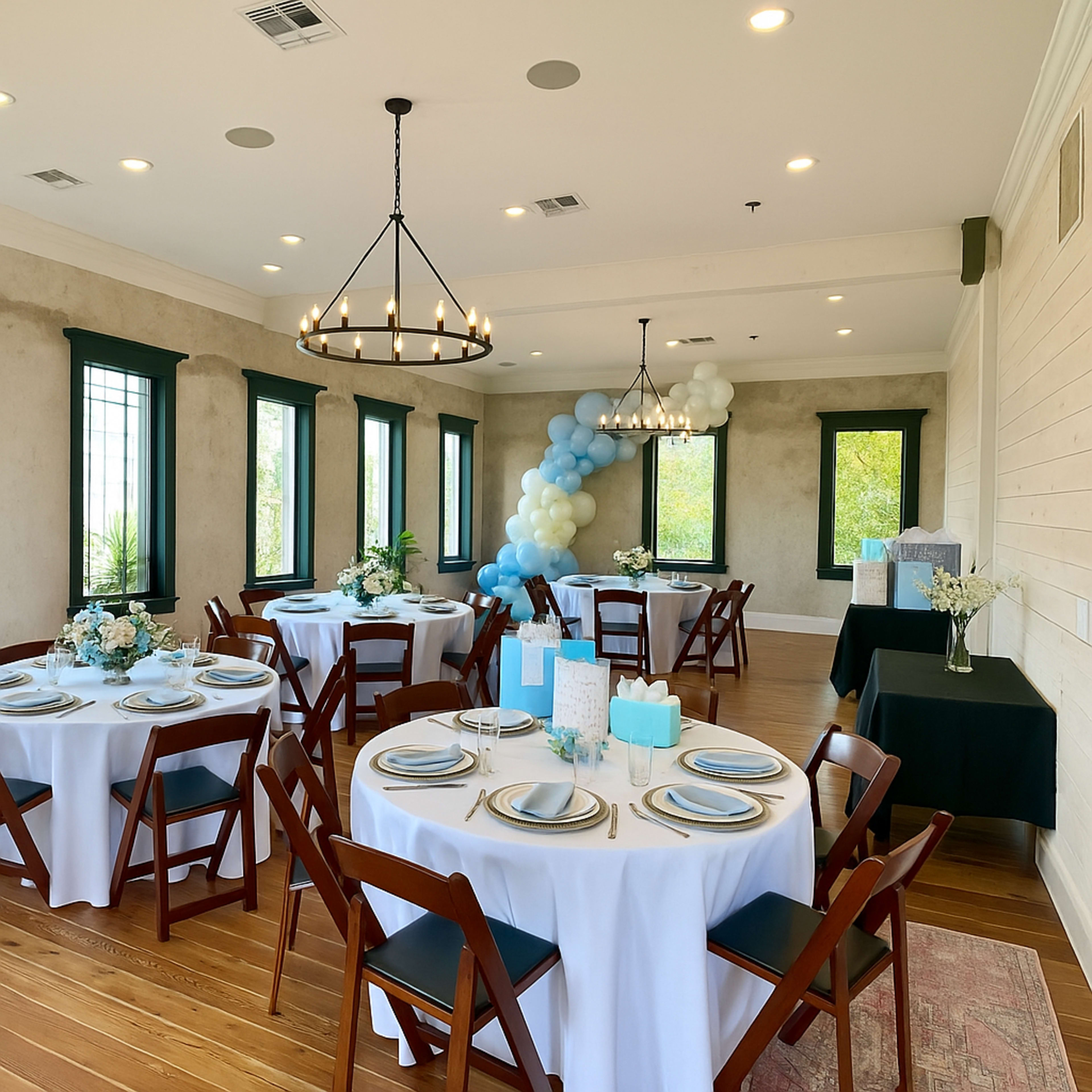 The image shows a well-decorated event room with round tables set for guests, featuring white tablecloths, elegant dishes, and a balloon arch in shades of blue and white.