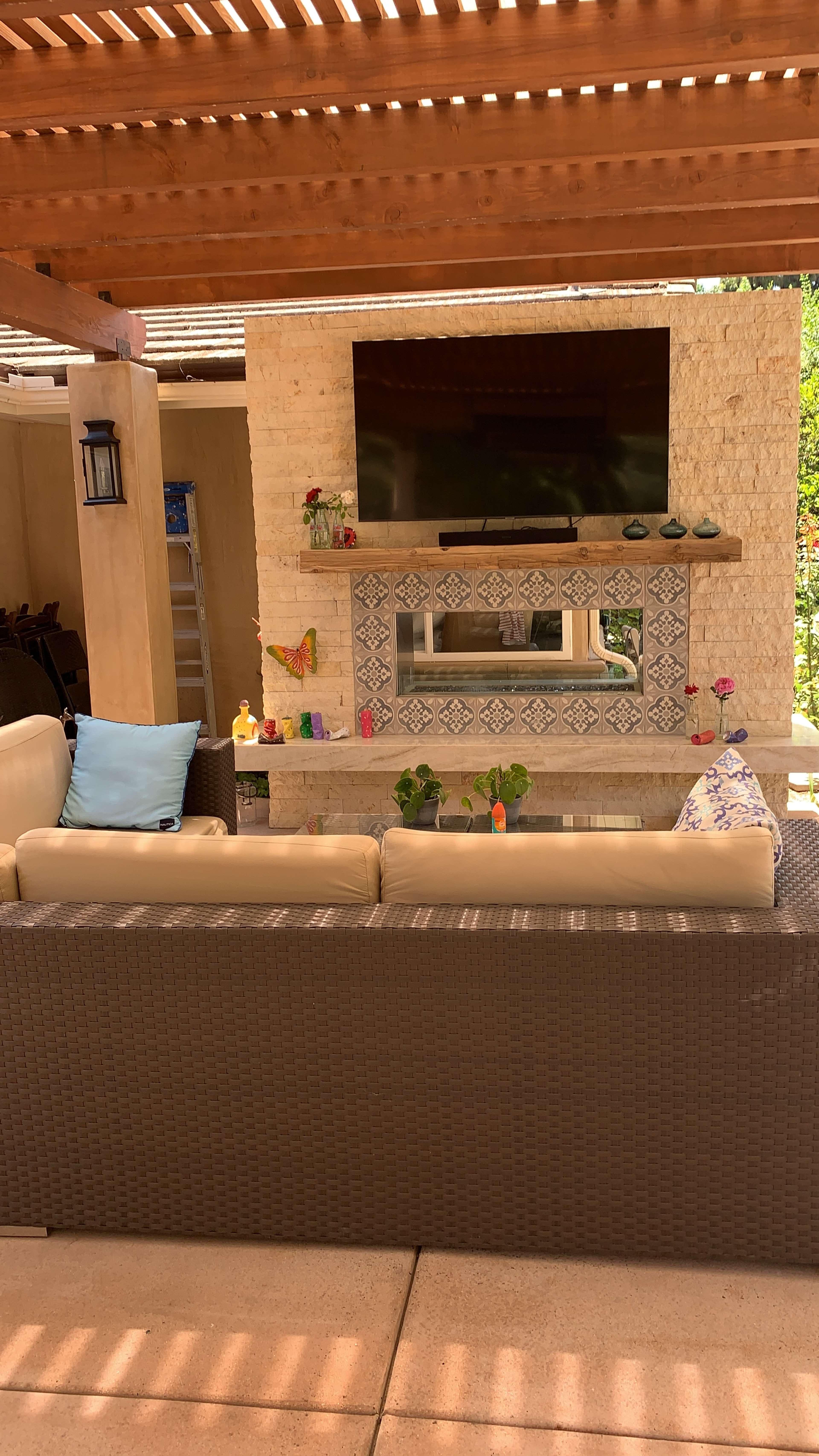 A covered outdoor seating area features a television mounted above a decorative stone fireplace, with potted plants and colorful decor on display.