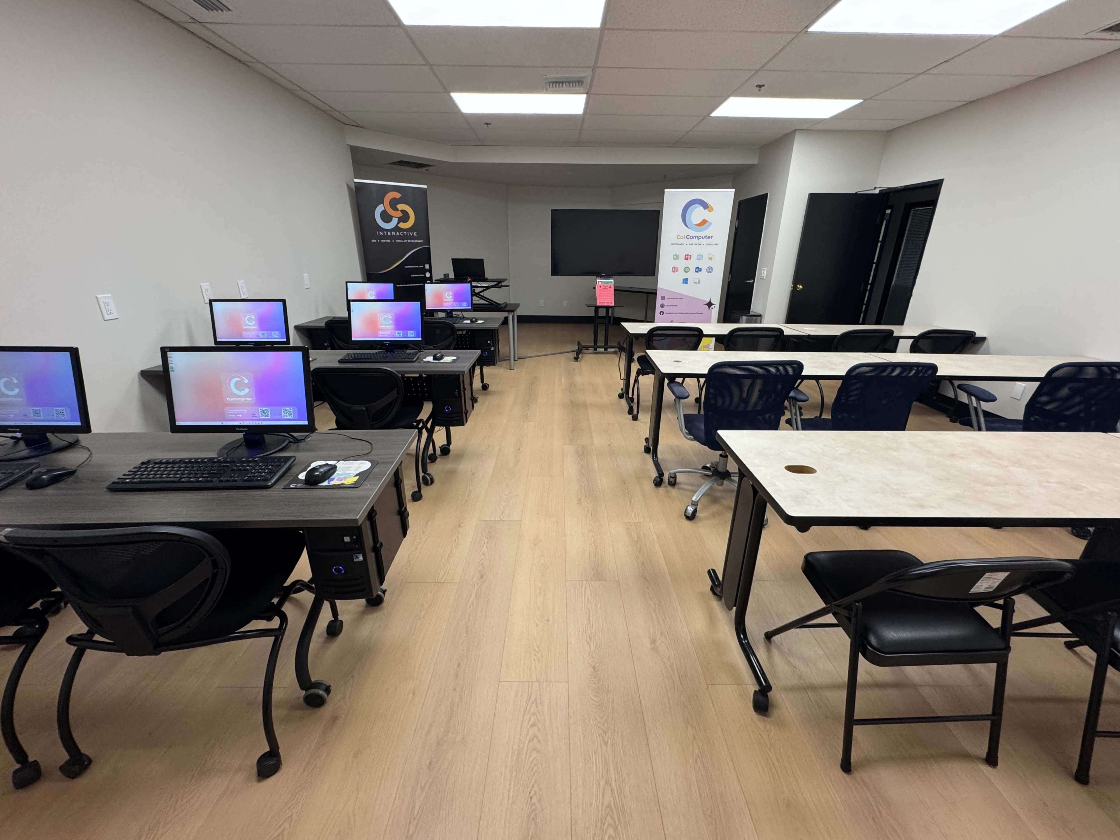The image shows a modern classroom with multiple computer workstations and tables arranged neatly, featuring promotional banners on the walls.
