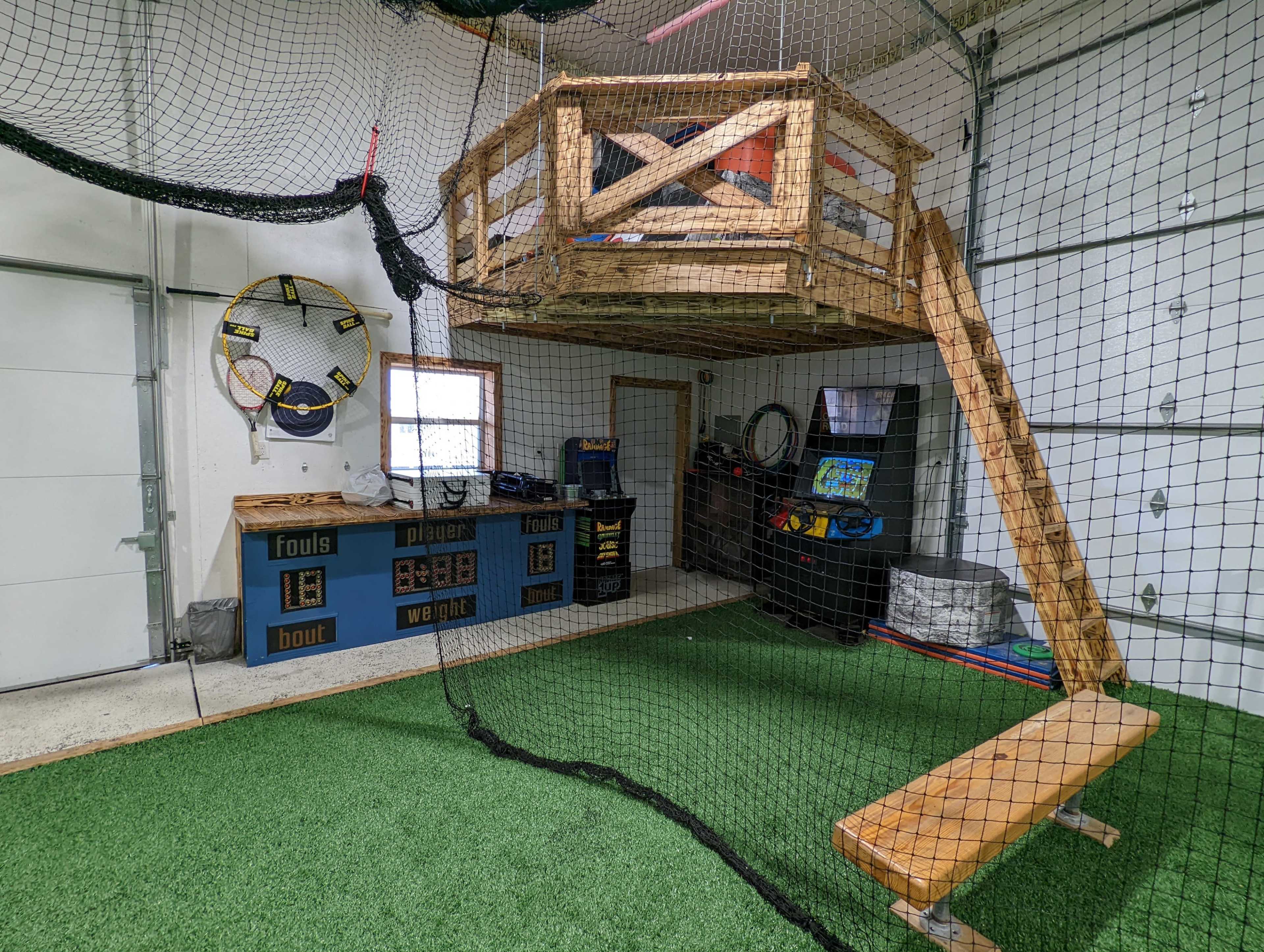 The image shows an indoor play area featuring a wooden platform elevated by netting, surrounded by artificial turf and various sports equipment.