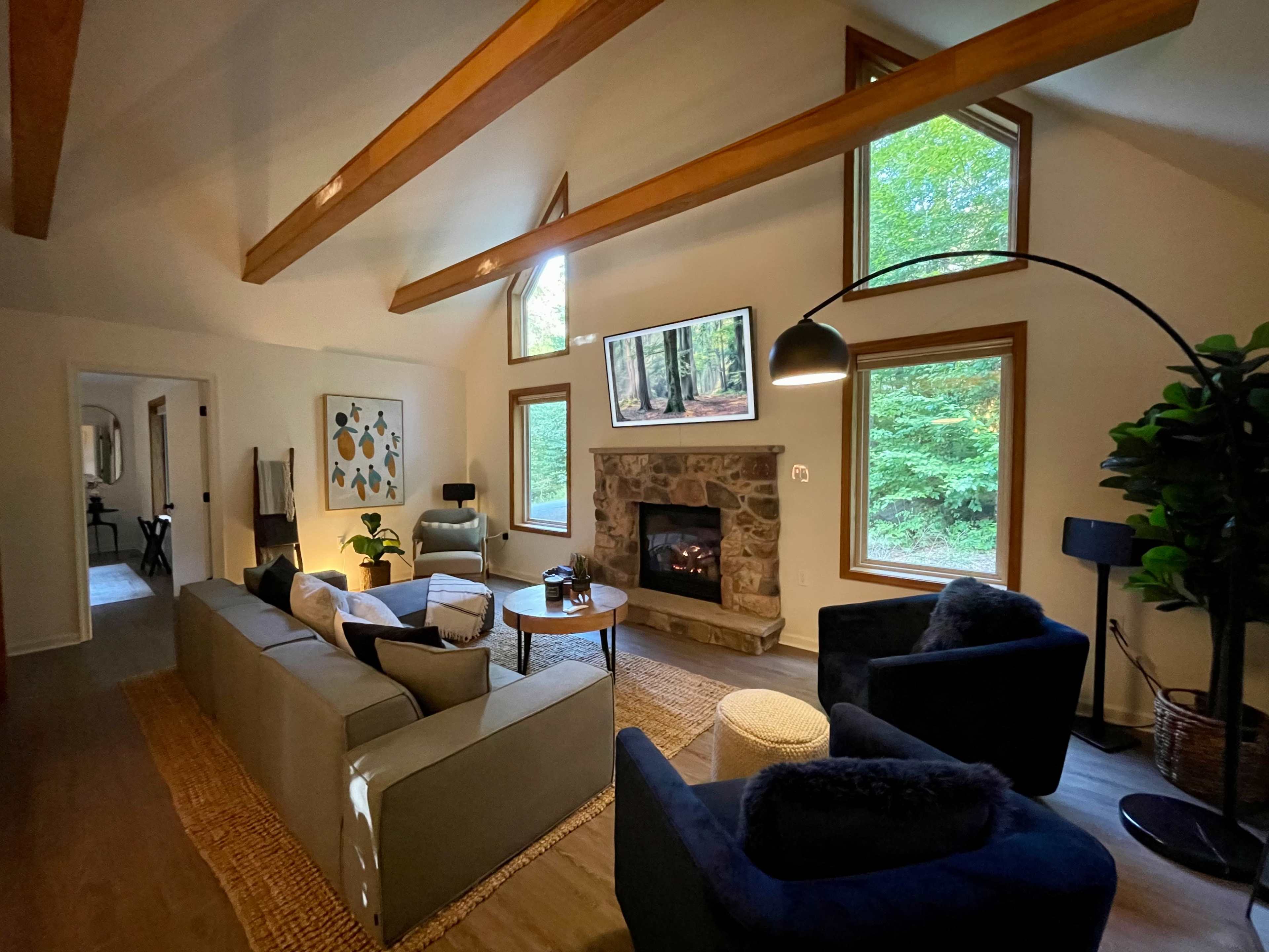 A cozy living room features a stone fireplace, large windows overlooking greenery, and modern furniture arrangements.