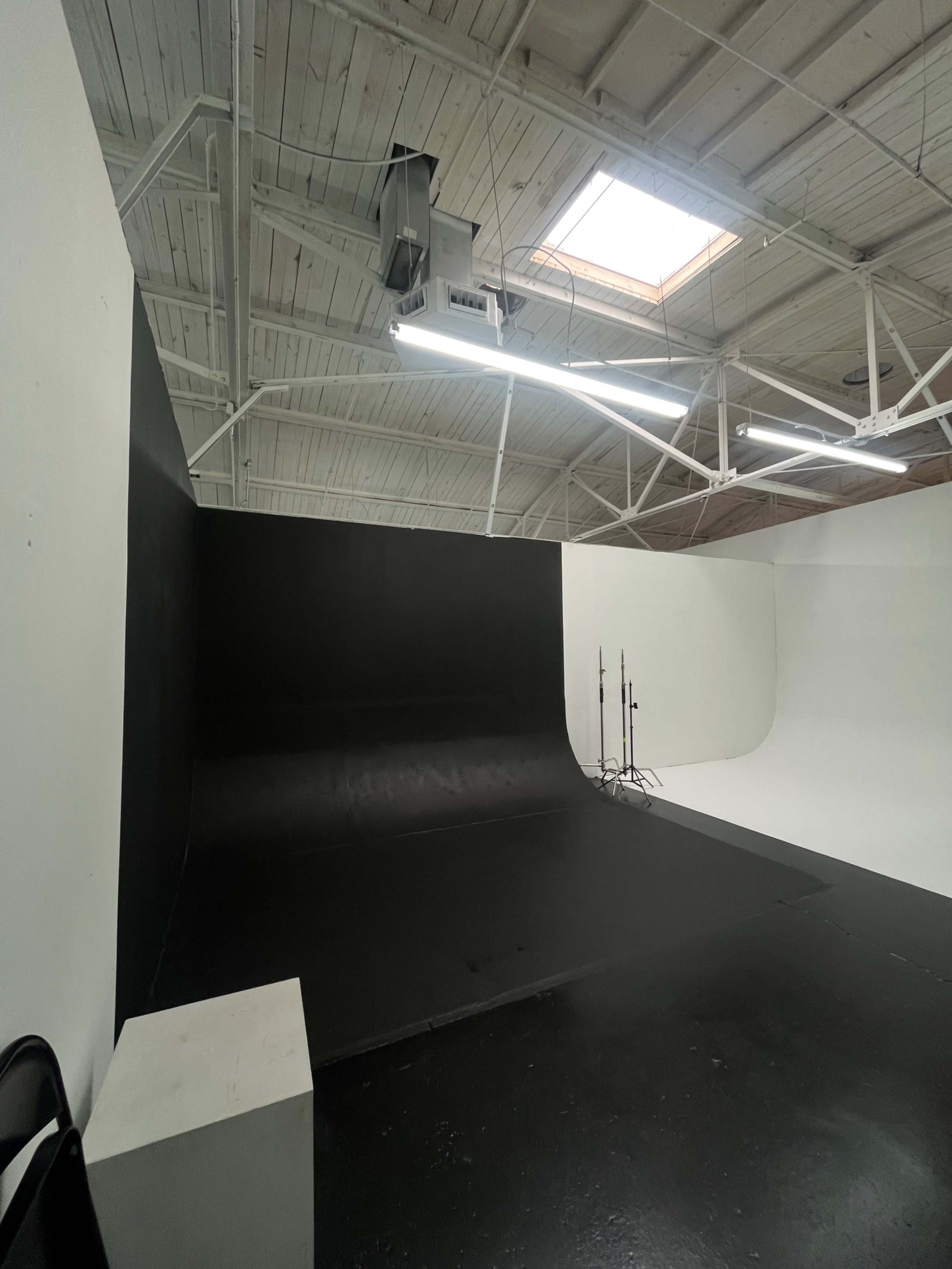 A photography studio features a large black backdrop and a white curved wall, with lighting fixtures mounted on the ceiling.