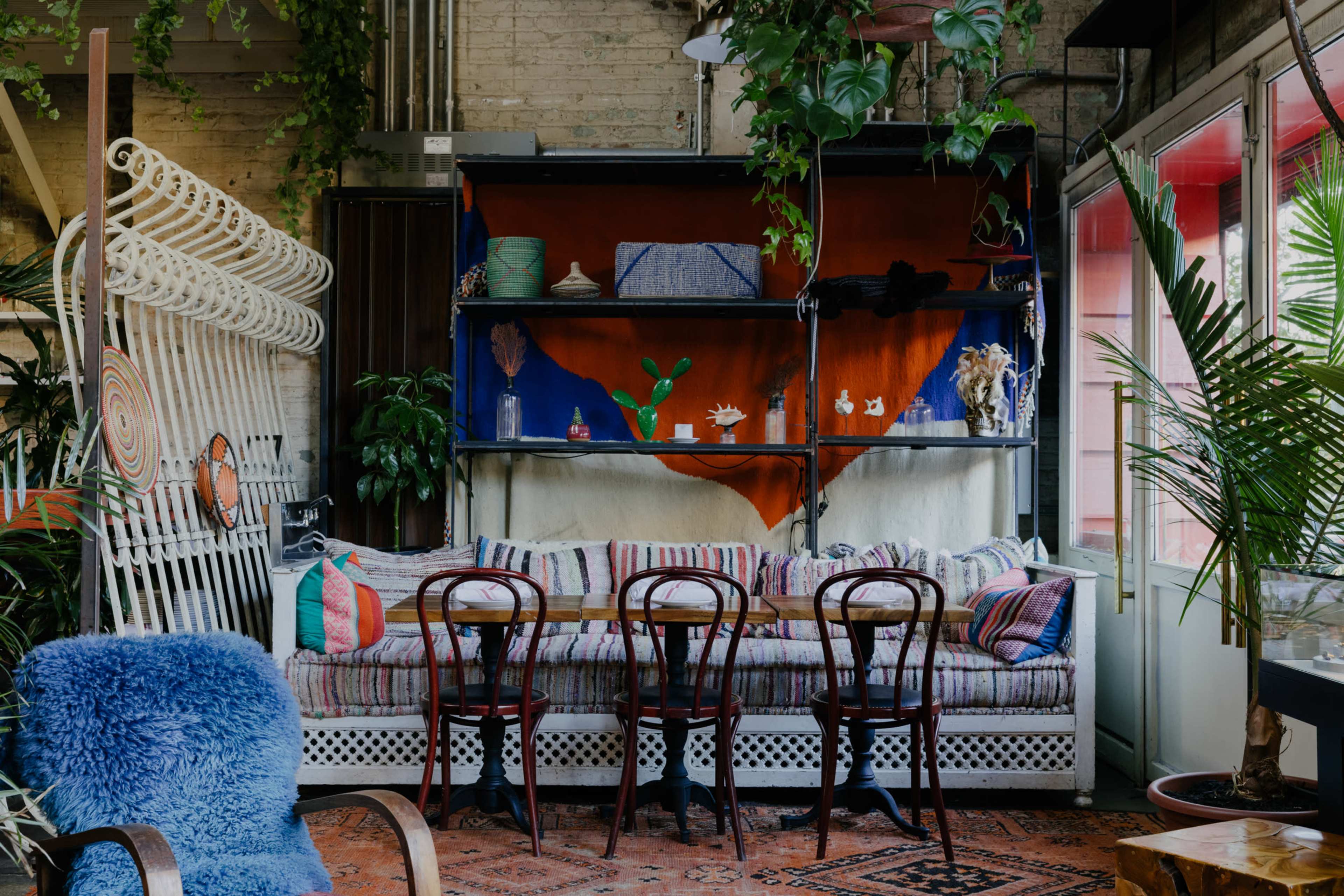 A cozy seating area with a patterned bench underneath a colorful shelf, surrounded by plants and wooden tables.