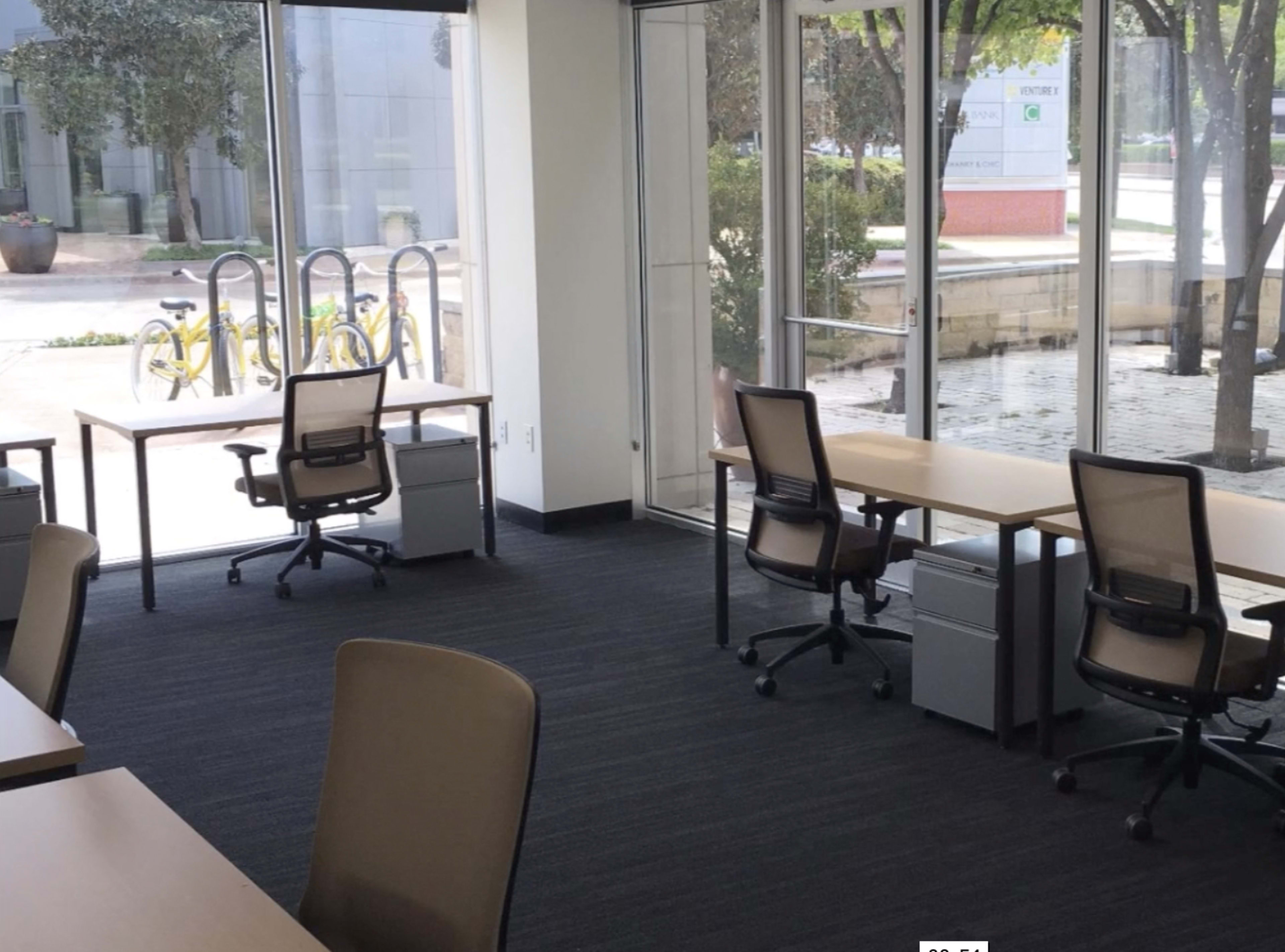 A modern office space with two desks and chairs beside large windows overlooking a courtyard.