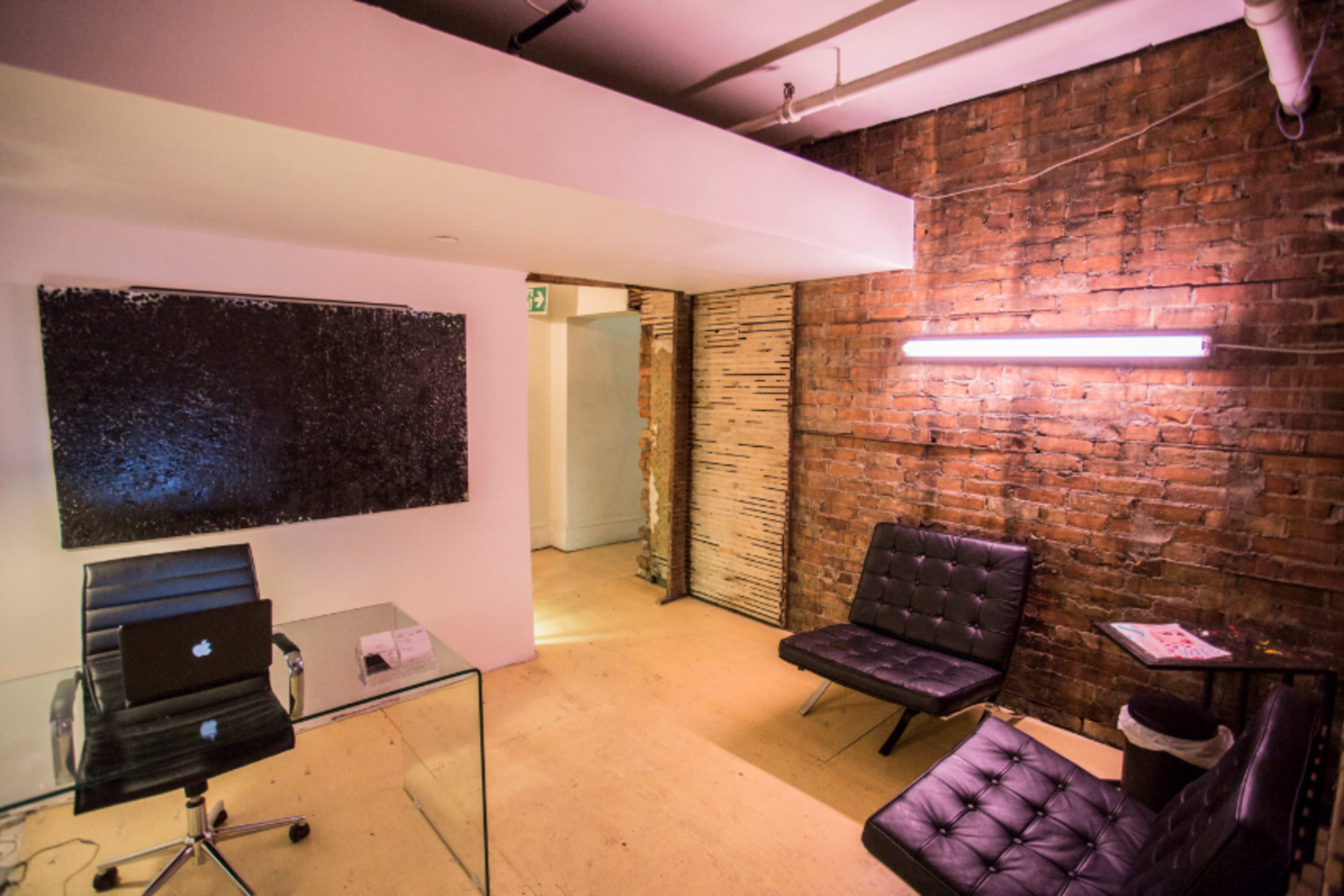 The image shows a minimalist office space with a glass desk, a black chair, and two black lounge chairs against a backdrop of exposed brick walls and sheeted wood.