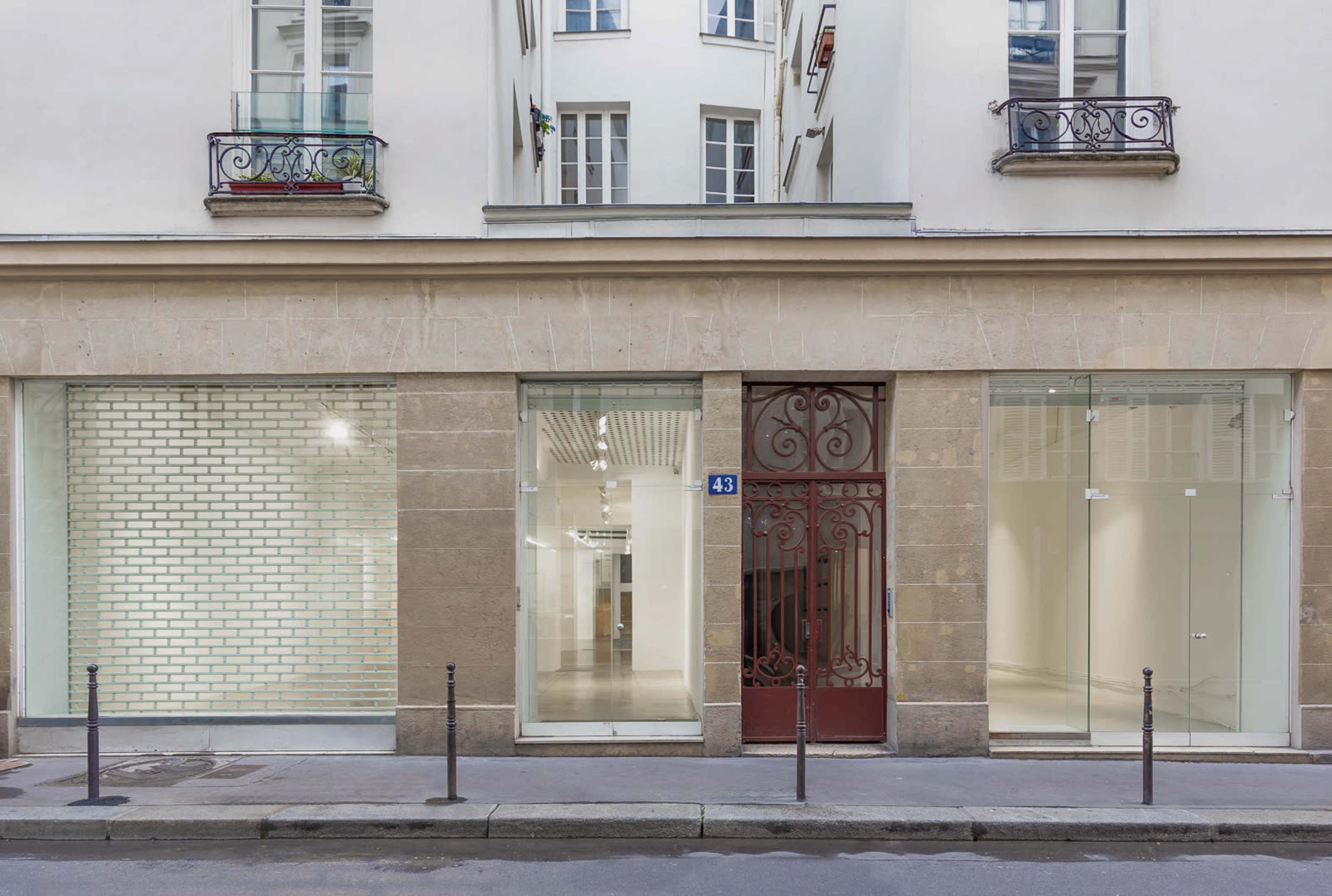 The image shows a storefront with large glass windows, a decorative metal gate, and a minimalist facade, located on a quiet Parisian street.