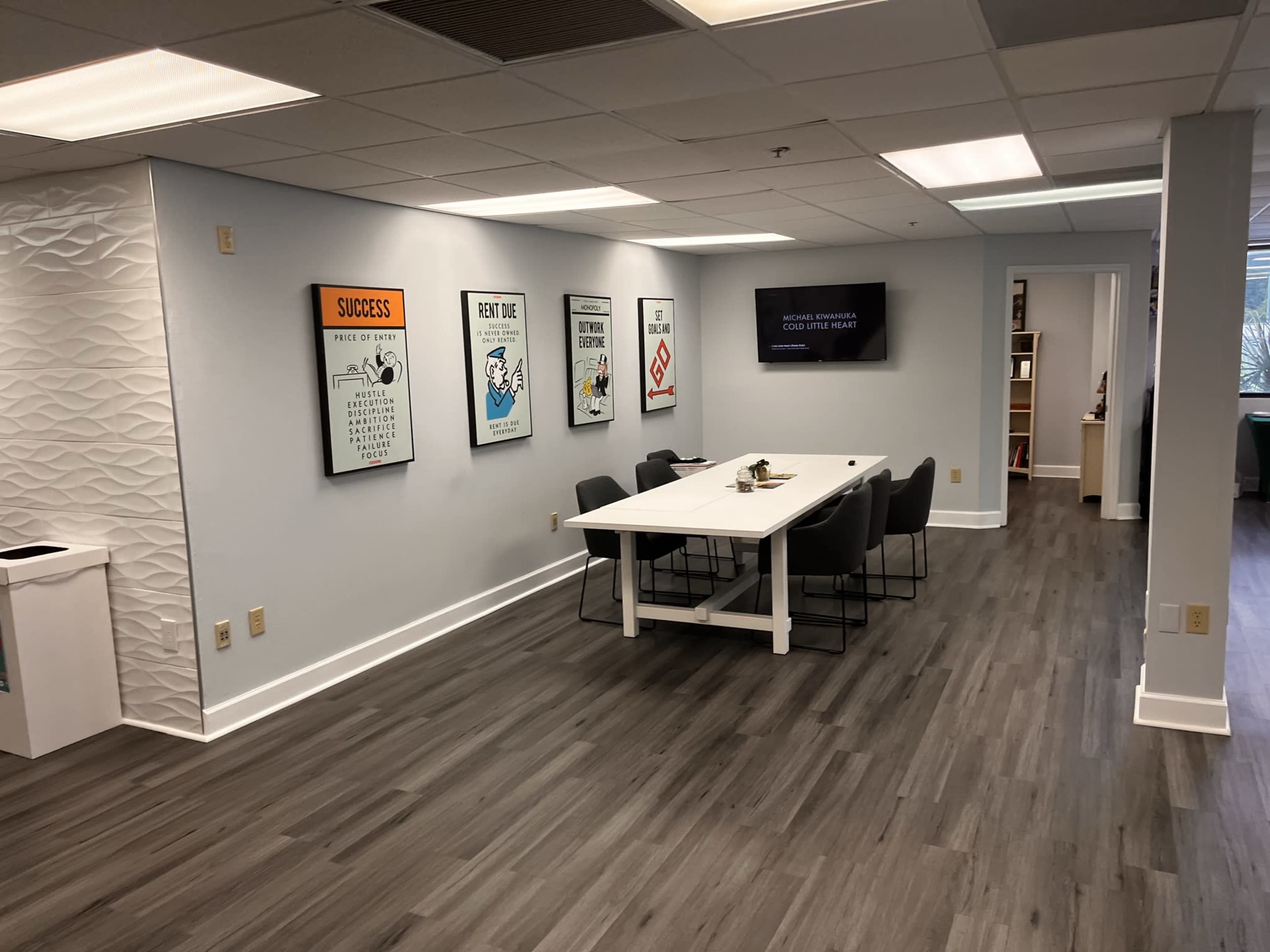 A modern office meeting room with a rectangular table surrounded by chairs, featuring wall art and a television on one side.