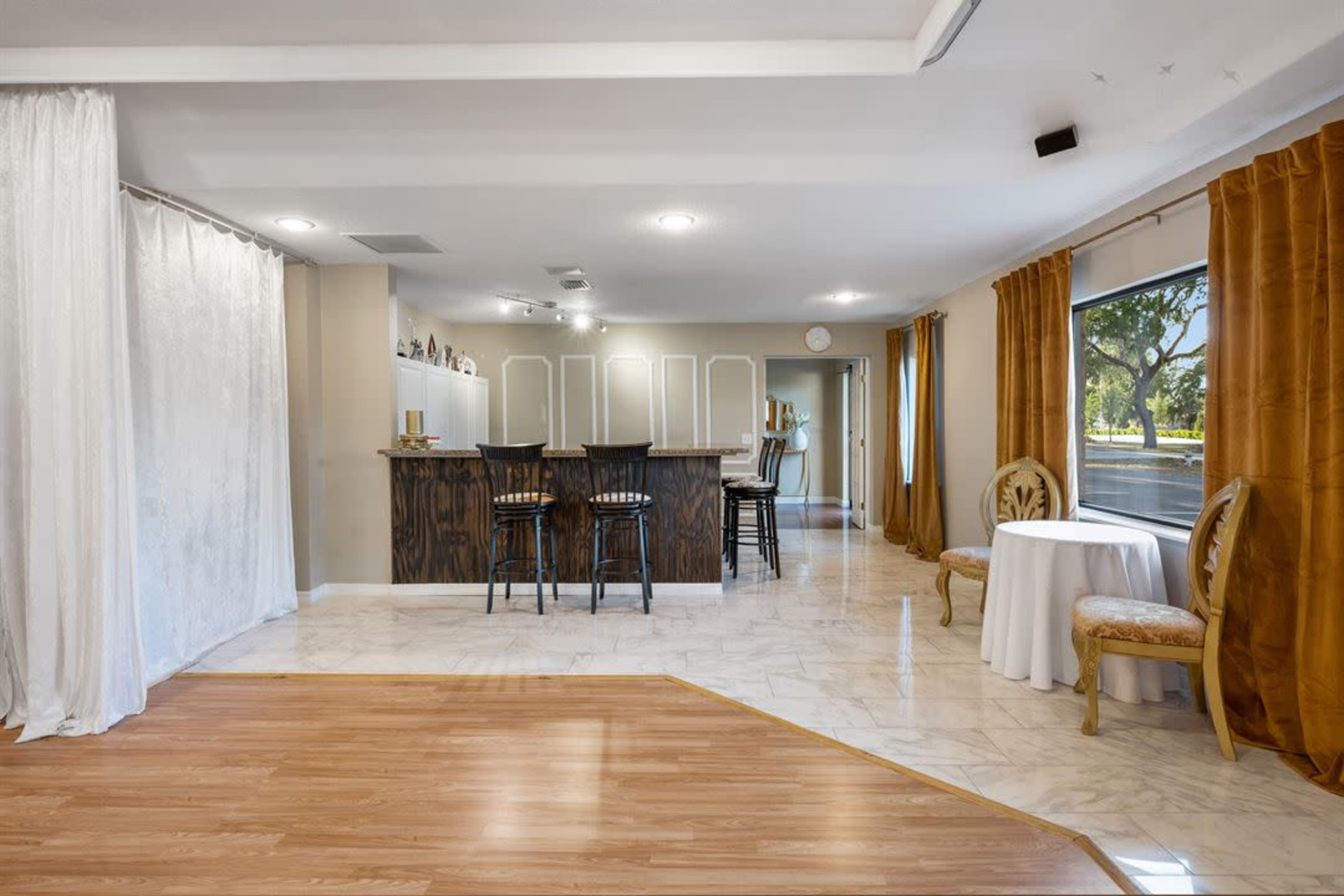 The image shows a modern interior space featuring a bar area with seating, a small table and chairs, and large windows with curtains.