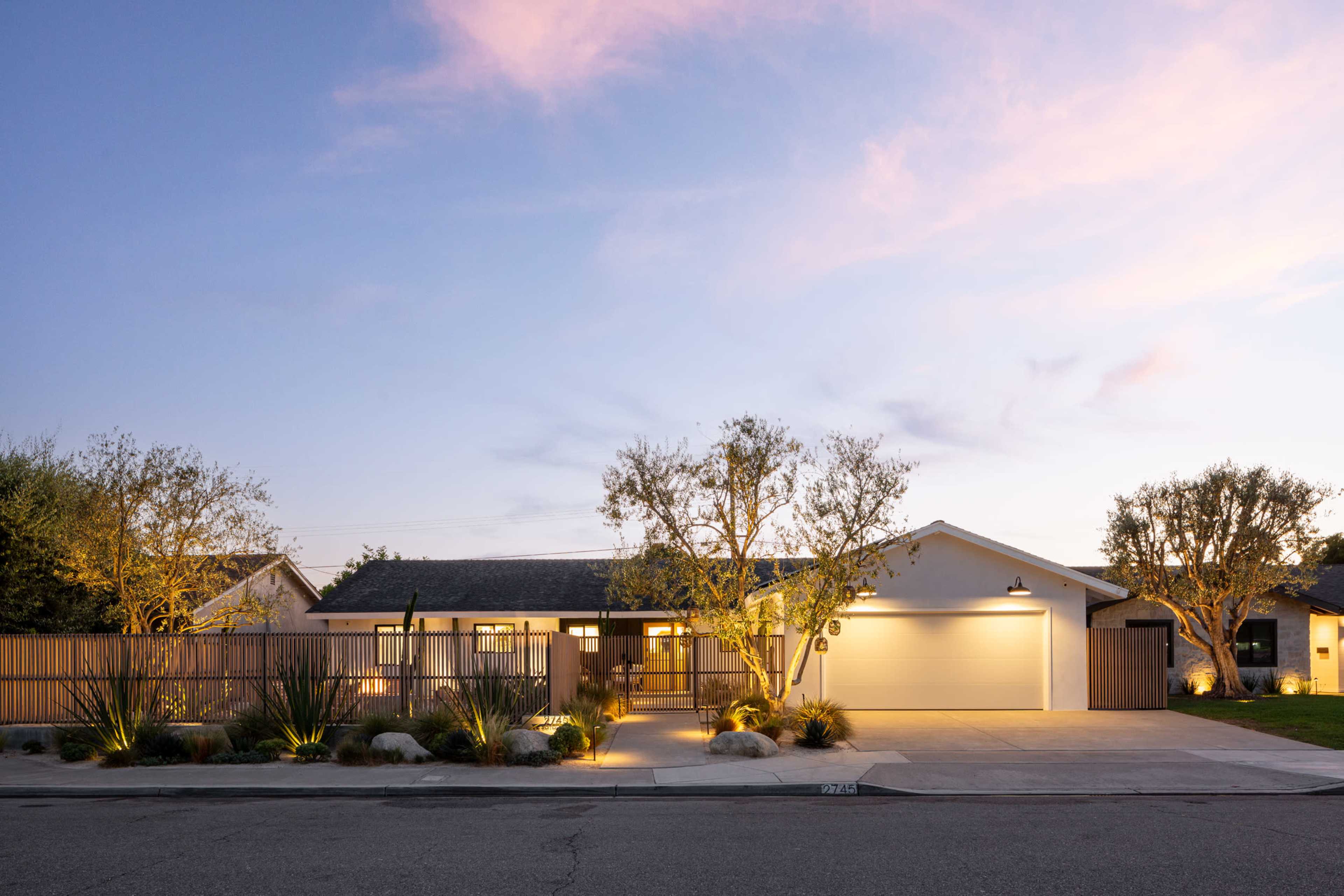 A contemporary single-story house with a landscaped front yard is illuminated under a twilight sky.