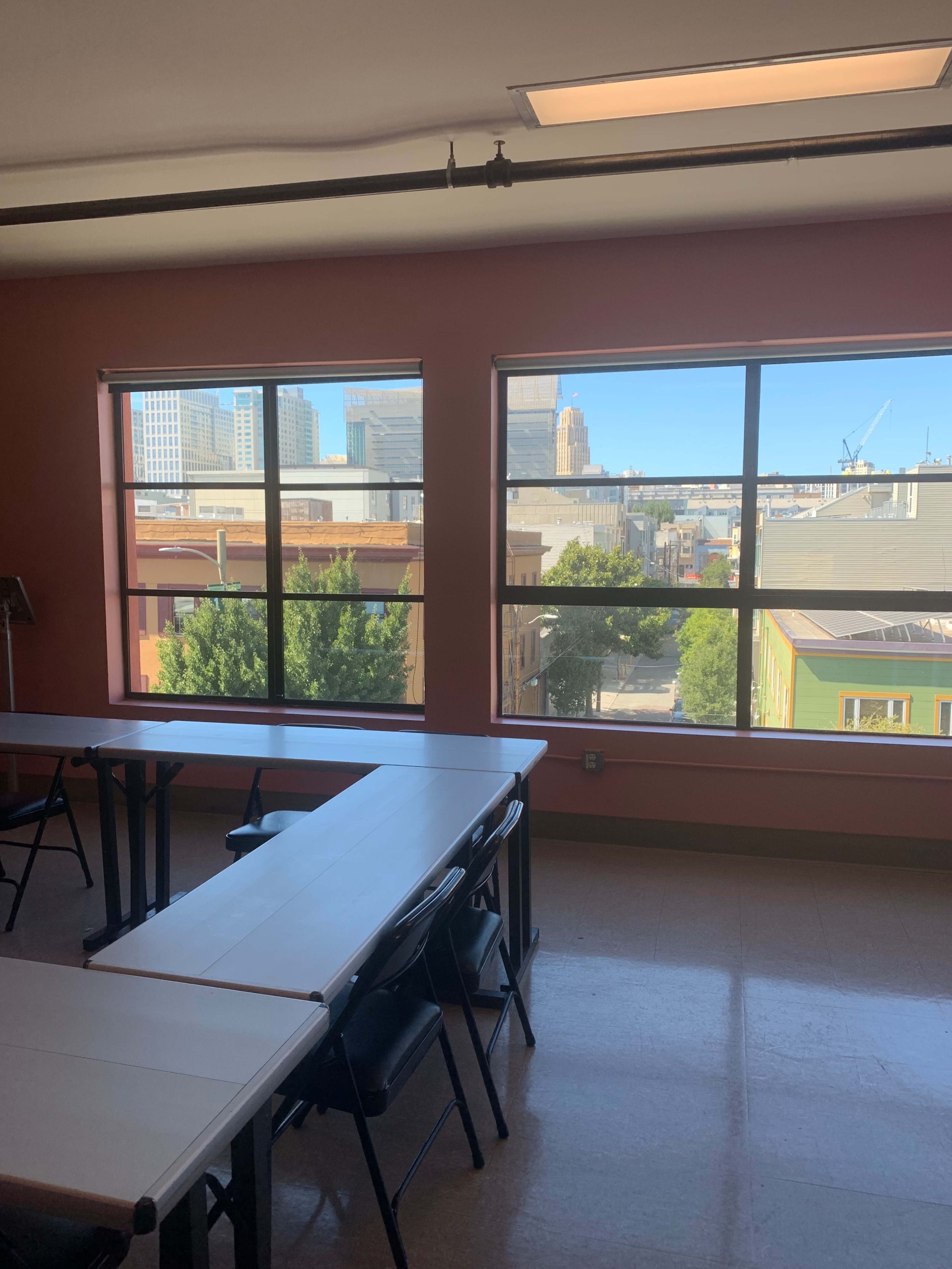 The image shows a classroom with long tables set up facing large windows that overlook a cityscape.