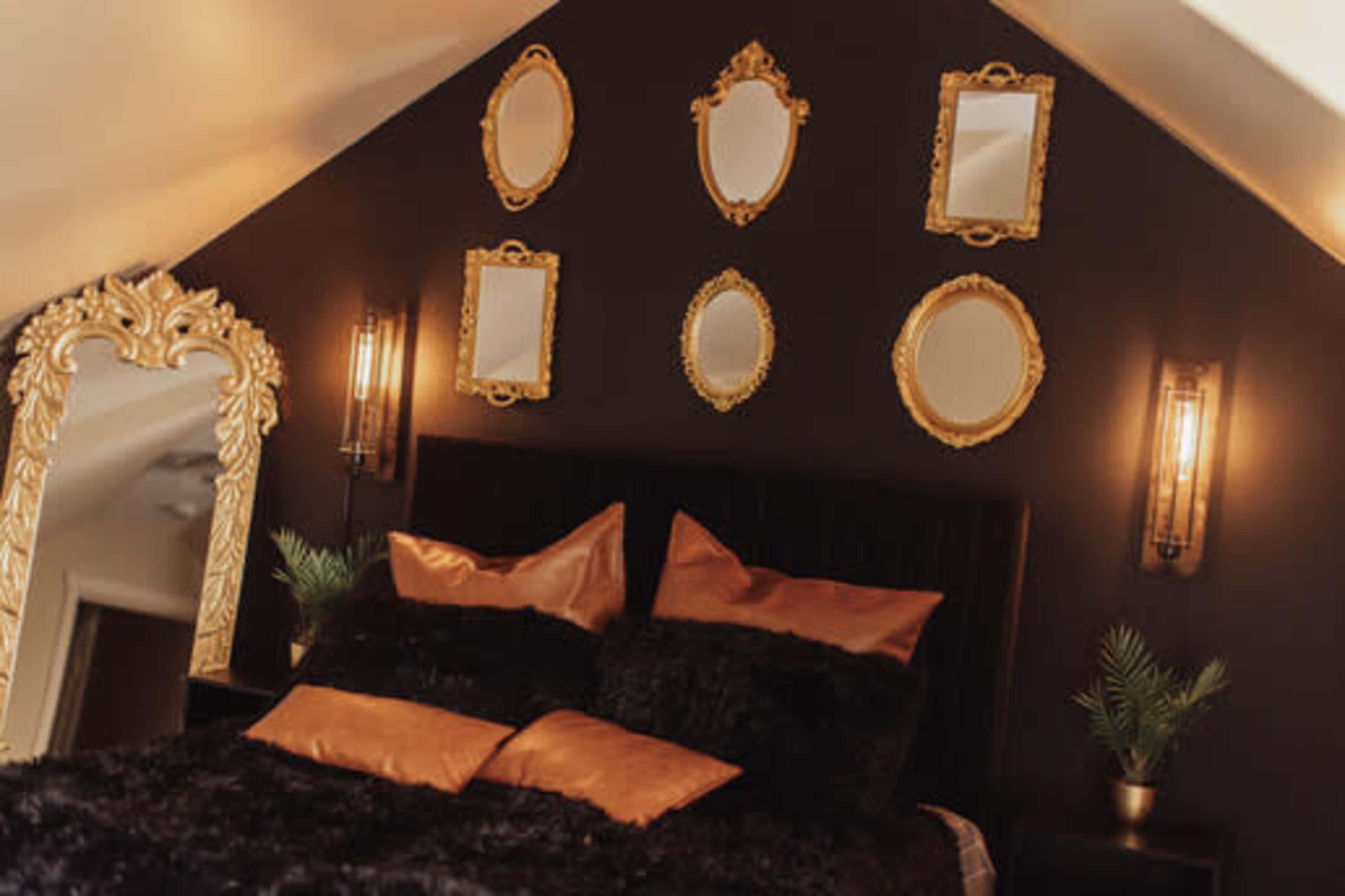 A dark-colored bedroom features a bed with black pillows and gold accents, complemented by ornate mirrors mounted on the wall and stylish wall sconces.