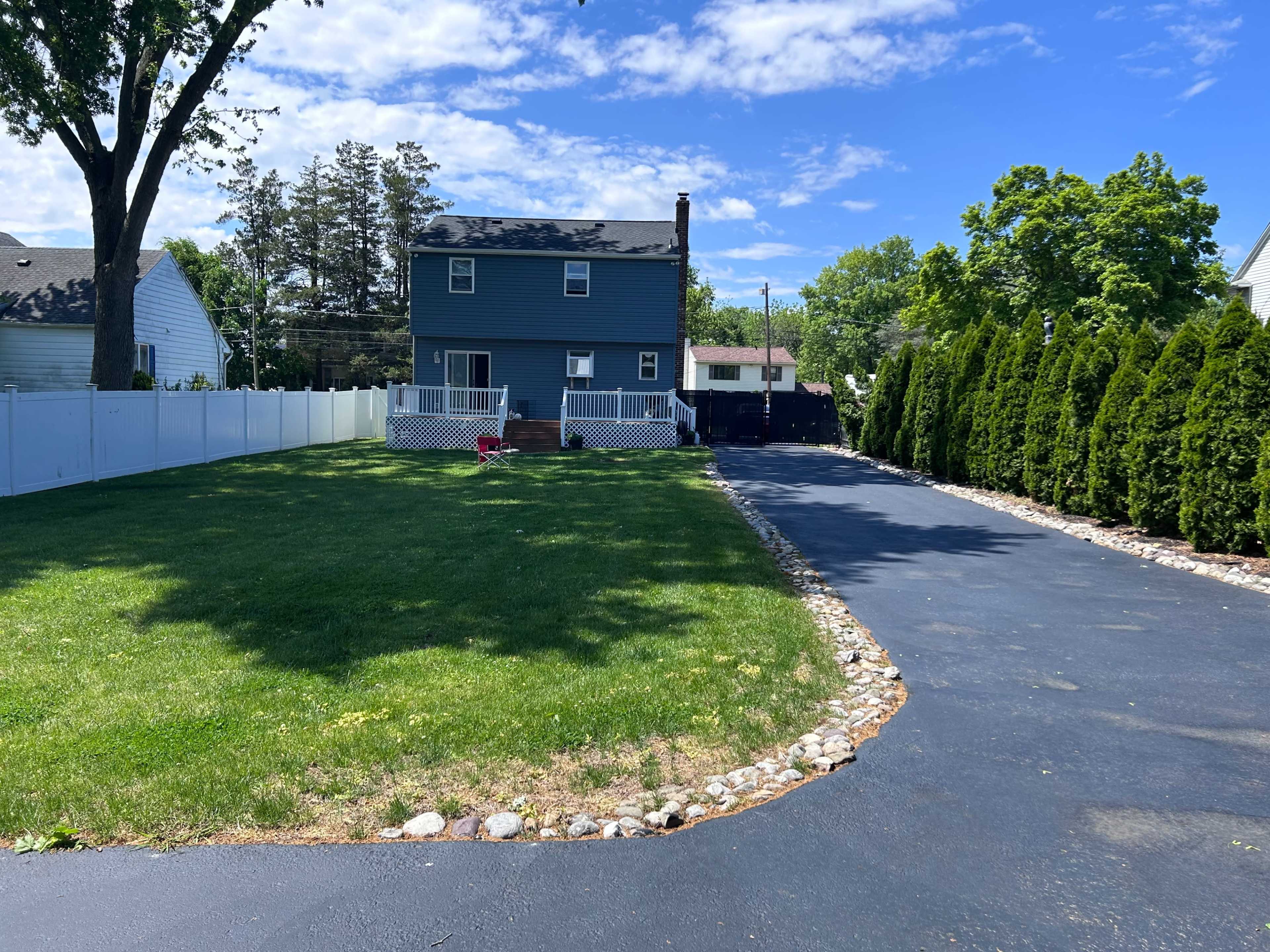 A blue two-story house with a white porch sits at the end of a paved driveway, surrounded by green grass and tall evergreen shrubs.