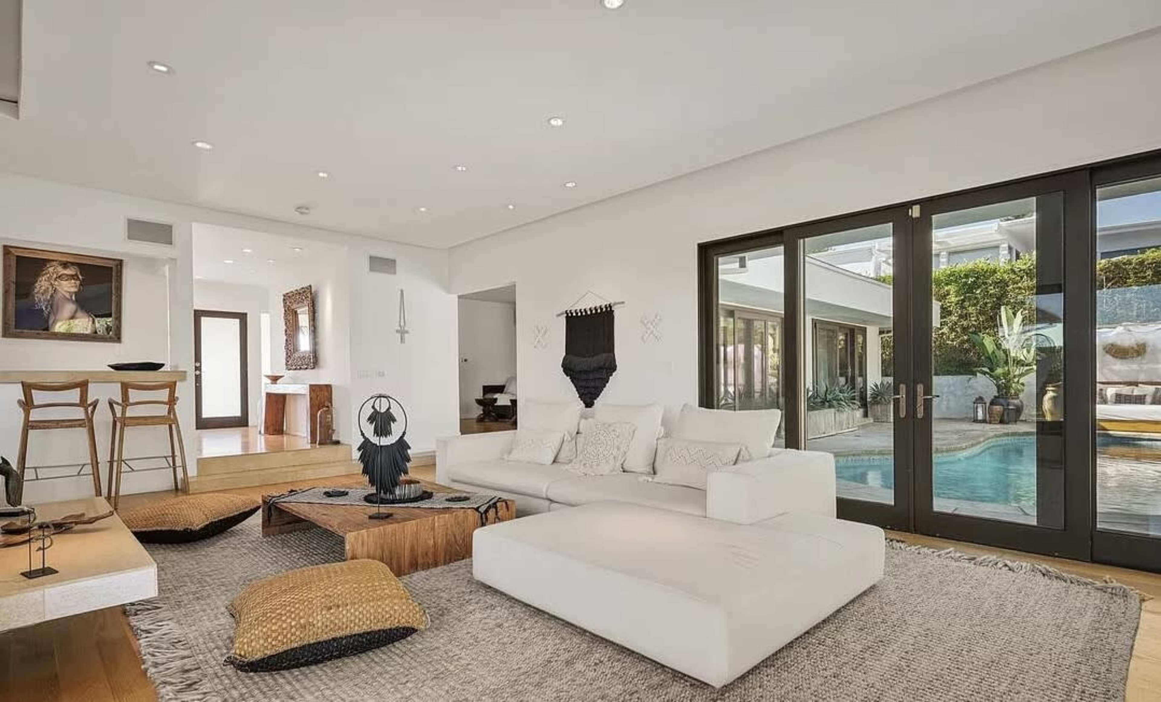 A modern living room with a white couch, wooden stools, and large glass doors leading to a pool area.