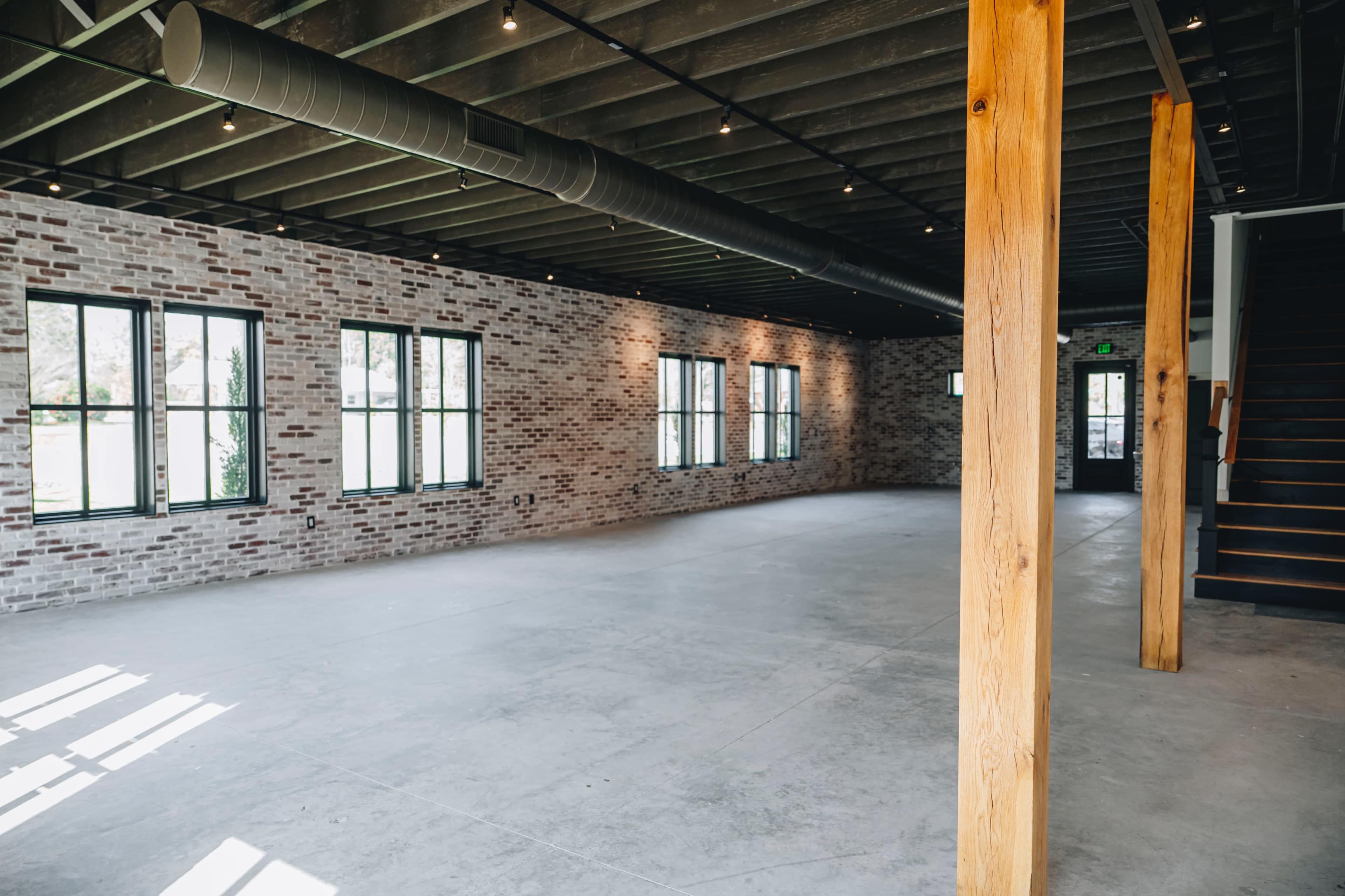 An empty industrial-style room with exposed brick walls, large windows, and wooden structural columns.