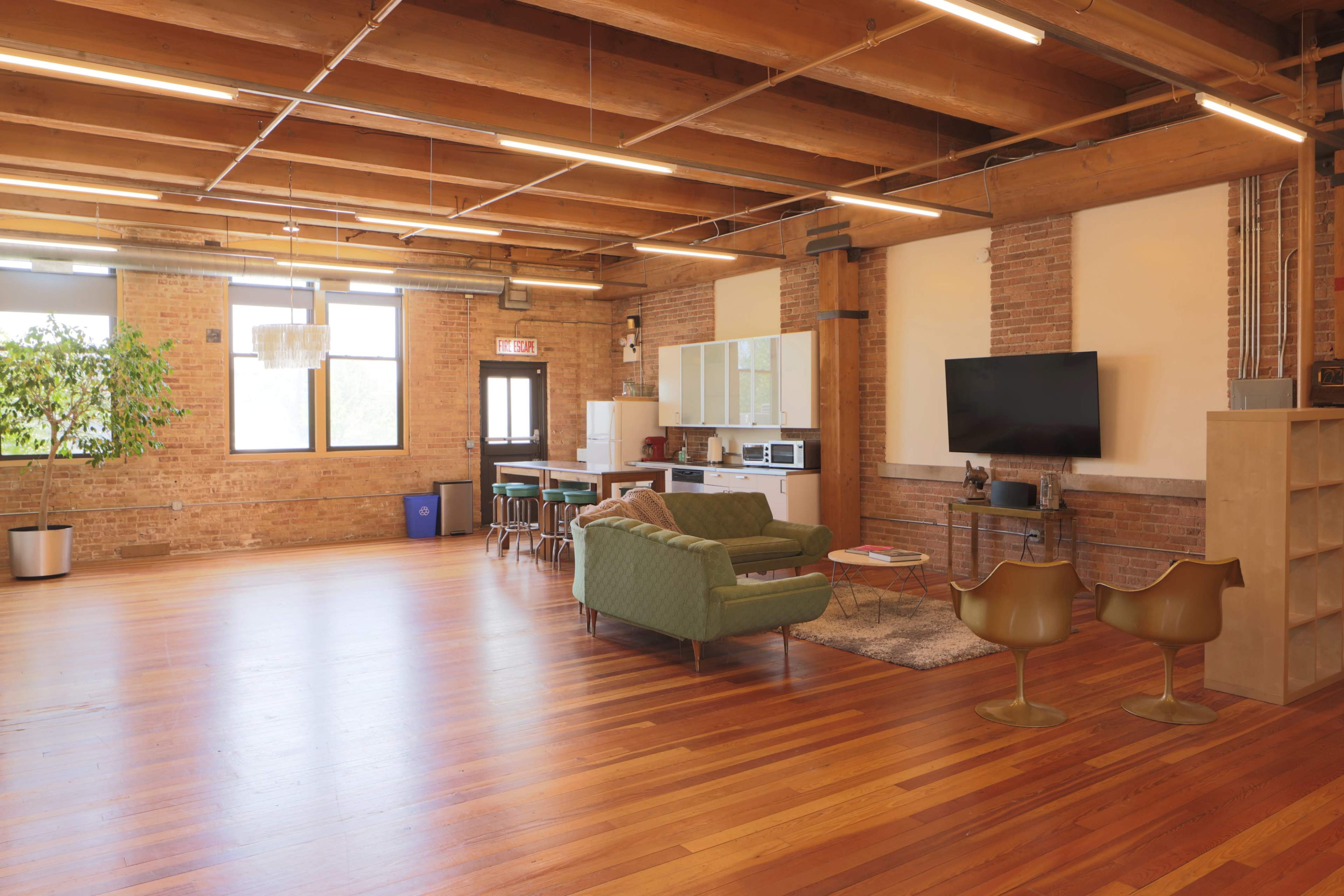 A spacious, modern loft with wooden beams, large windows, a living area featuring a green couch and two gold chairs, and a kitchen area against a brick wall.