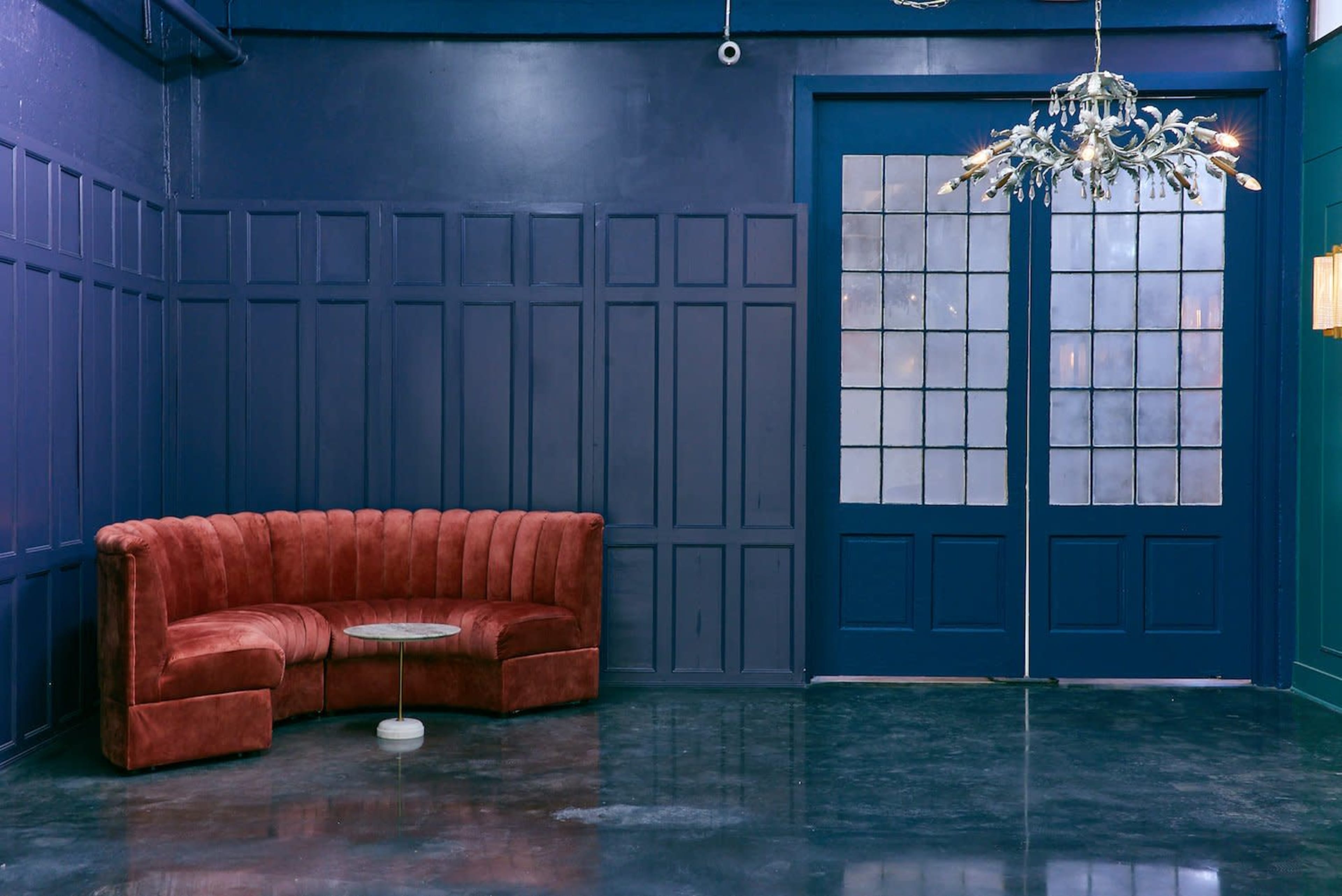 The image shows a corner seating area with a curved red velvet sofa and a glass-top coffee table against a dark blue wall featuring large glass double doors.