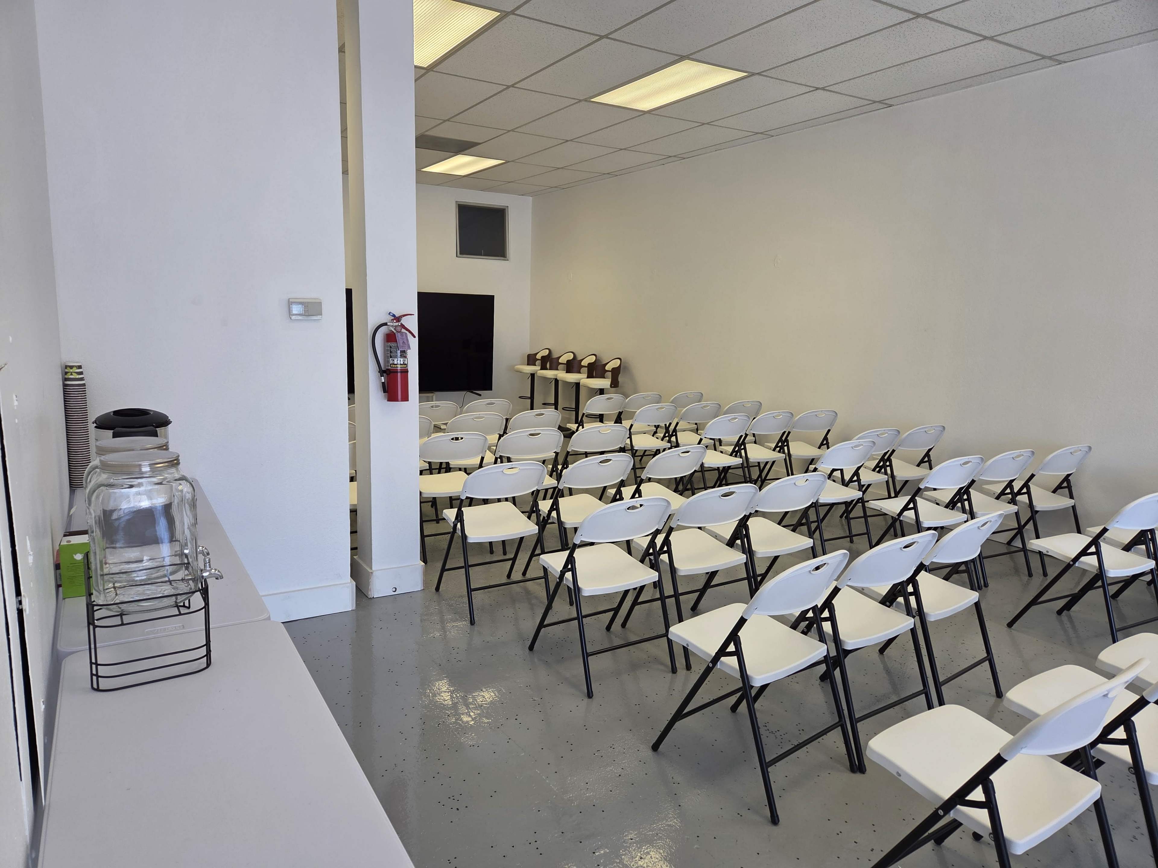 The image shows a spacious, empty room arranged with rows of white folding chairs facing a wall with a television and several coffee machines set up nearby.