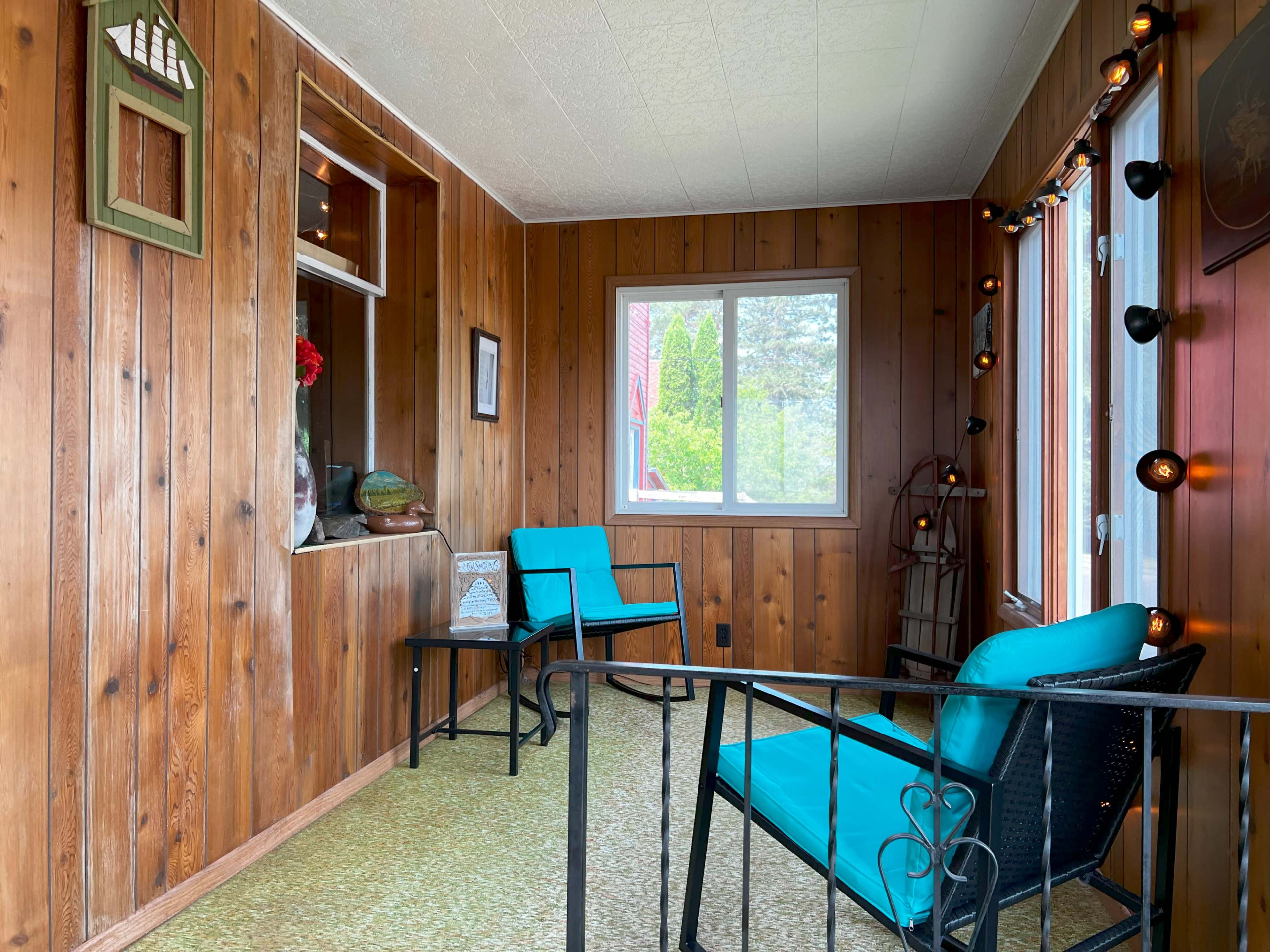 The image shows a sunroom with wooden paneling, two turquoise chairs, and a window overlooking greenery.