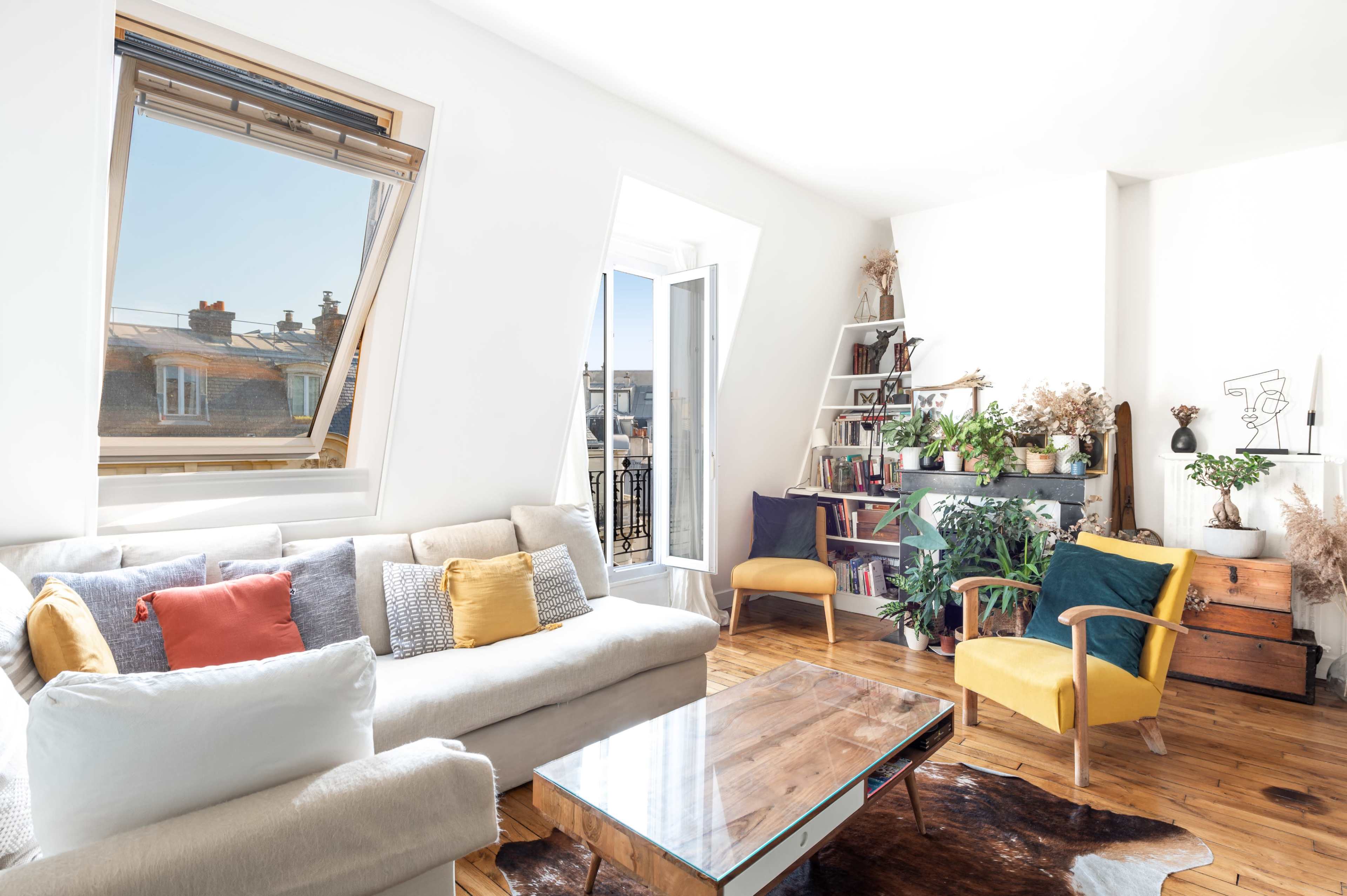 A bright living room featuring a light-colored sofa, a wooden coffee table, and a bookshelf filled with plants and decor, complemented by an open window with a view of rooftops.