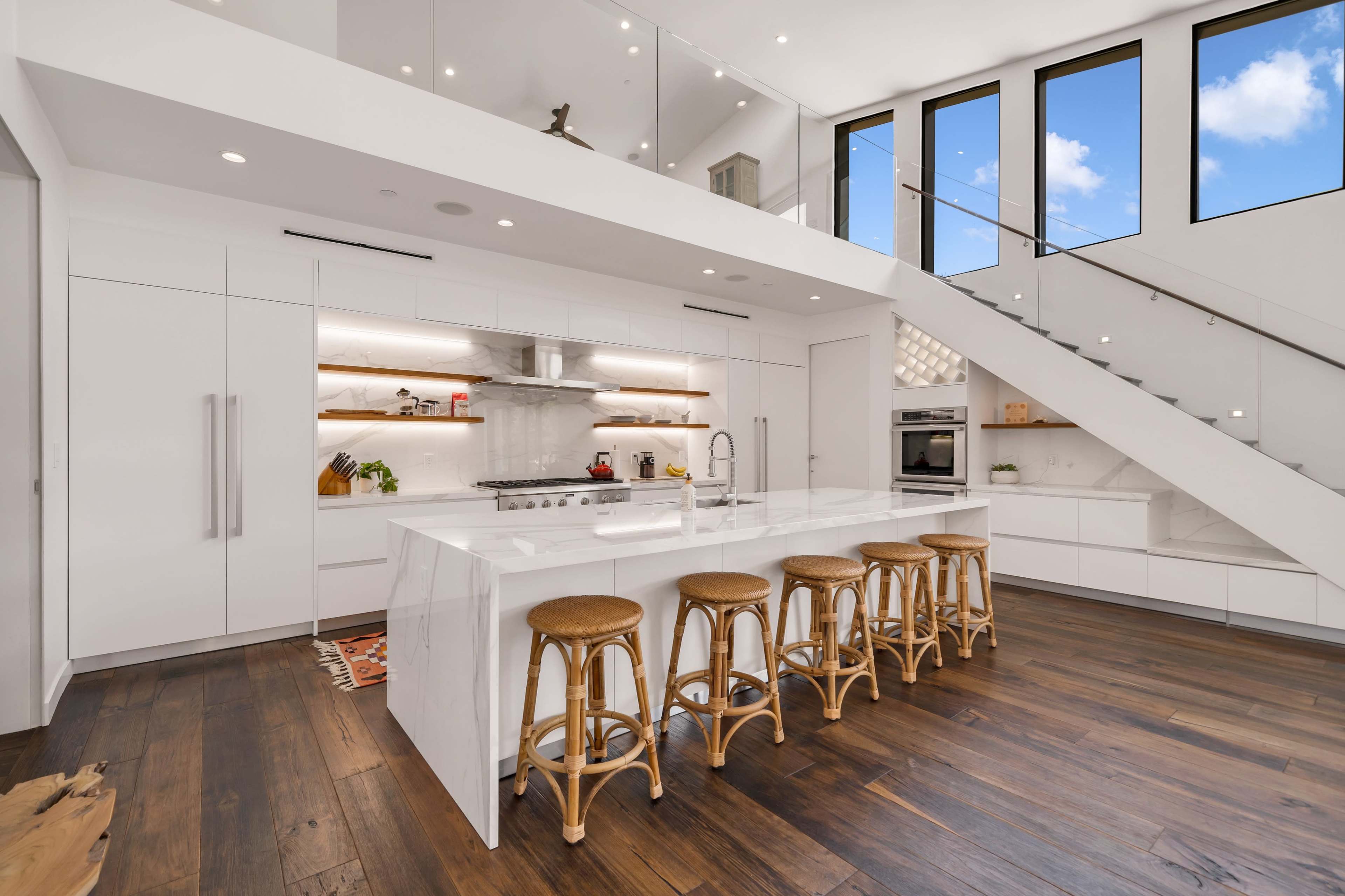 The kitchen features white cabinetry, a large marble island with seating, and wooden floors under ample natural light streaming through tall windows.