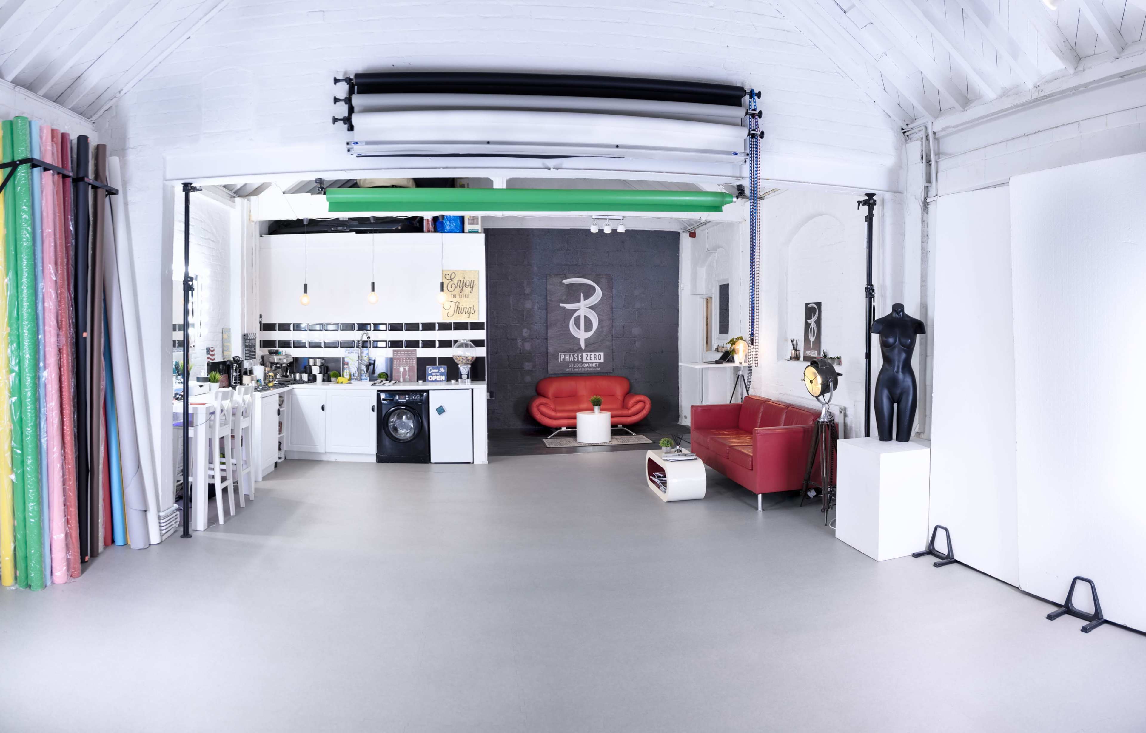 A spacious, white-walled studio featuring a kitchen area, a red sofa, a black mannequin, and various colorful backdrops along one wall.