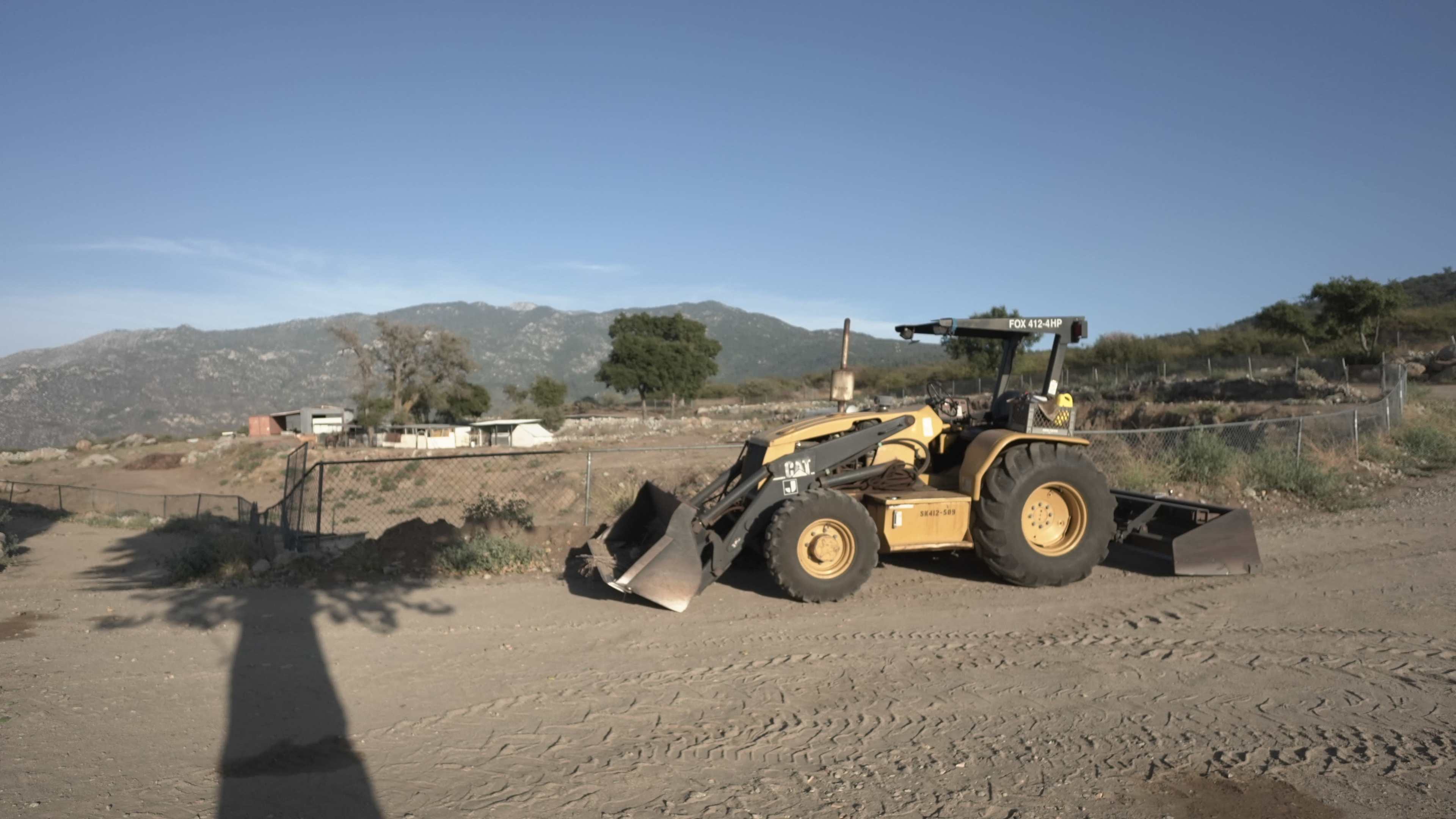 A yellow front-end loader is parked on a dirt lot with a mountainous landscape in the background.