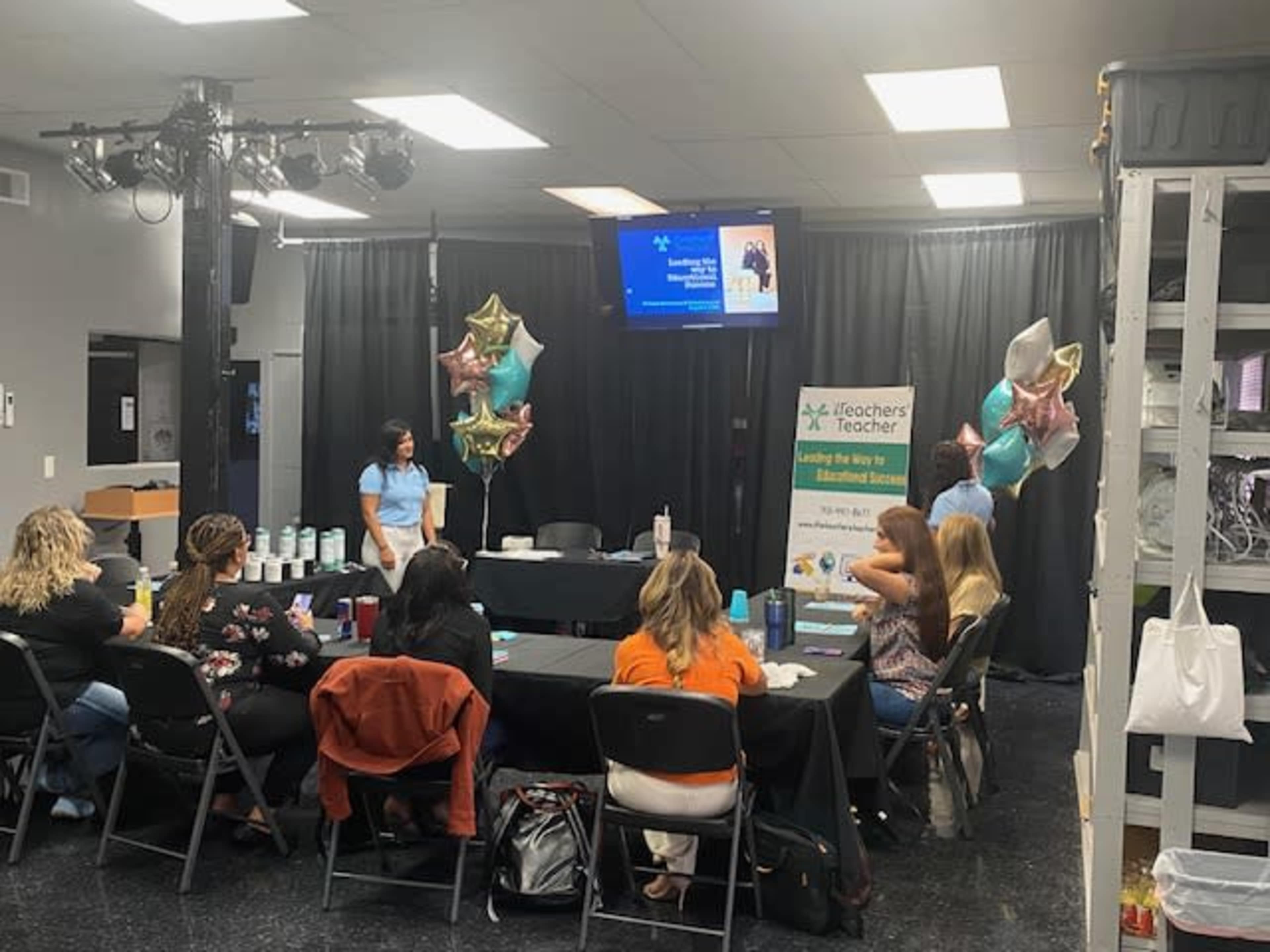 A group of people sits at a long black table during a presentation, while balloons and a banner promoting iTeachers are displayed in the background.