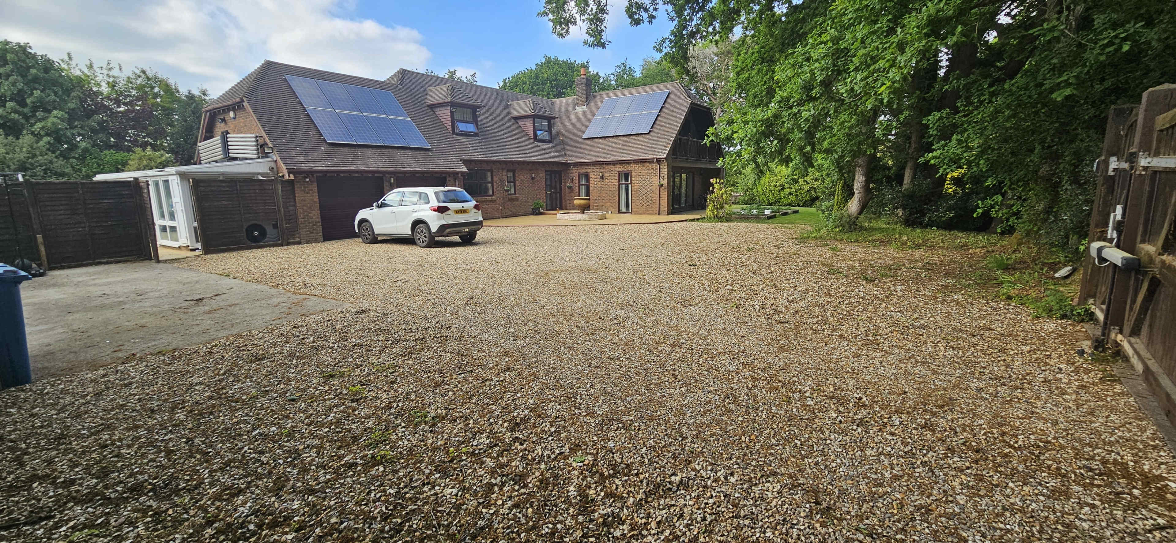 A large house with solar panels sits on a gravel driveway surrounded by trees.