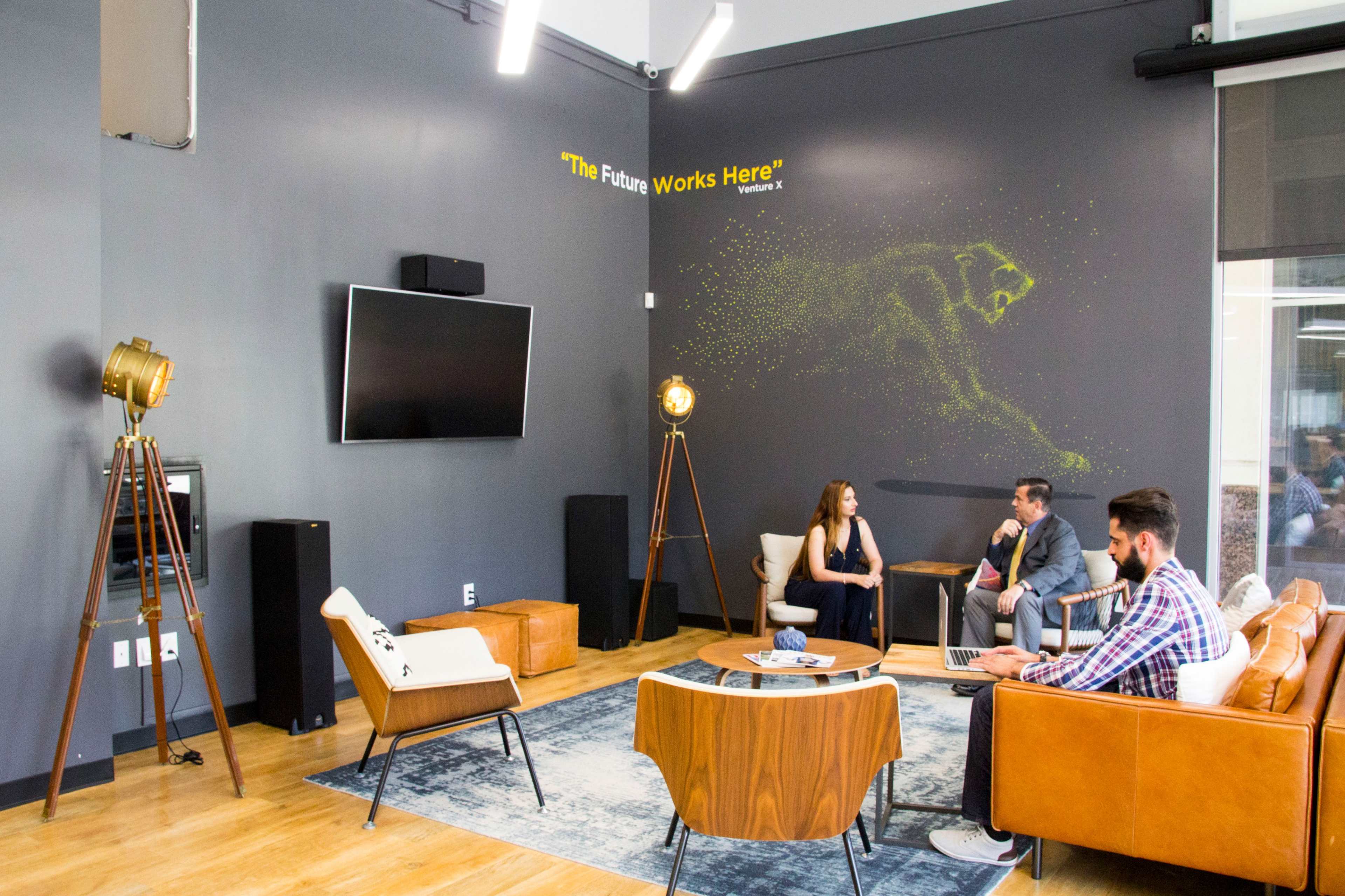 A modern office space features a seating area with three people engaged in conversation, a large television, and decorative lighting on a dark wall with the phrase "The Future Works Here."