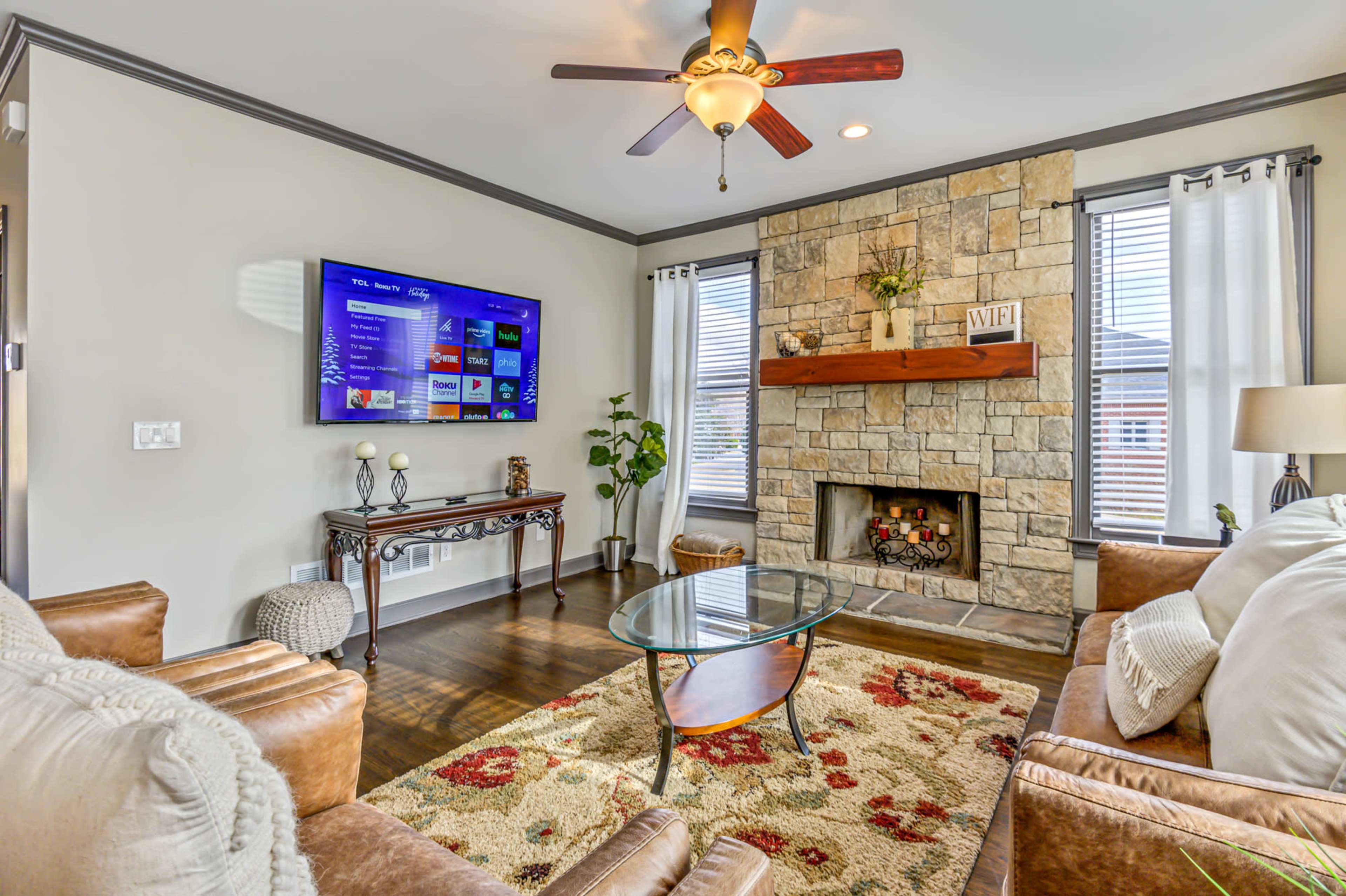 The living room features a stone fireplace, a flat-screen TV mounted on the wall, a glass coffee table, and a sofa set arranged around a patterned area rug.