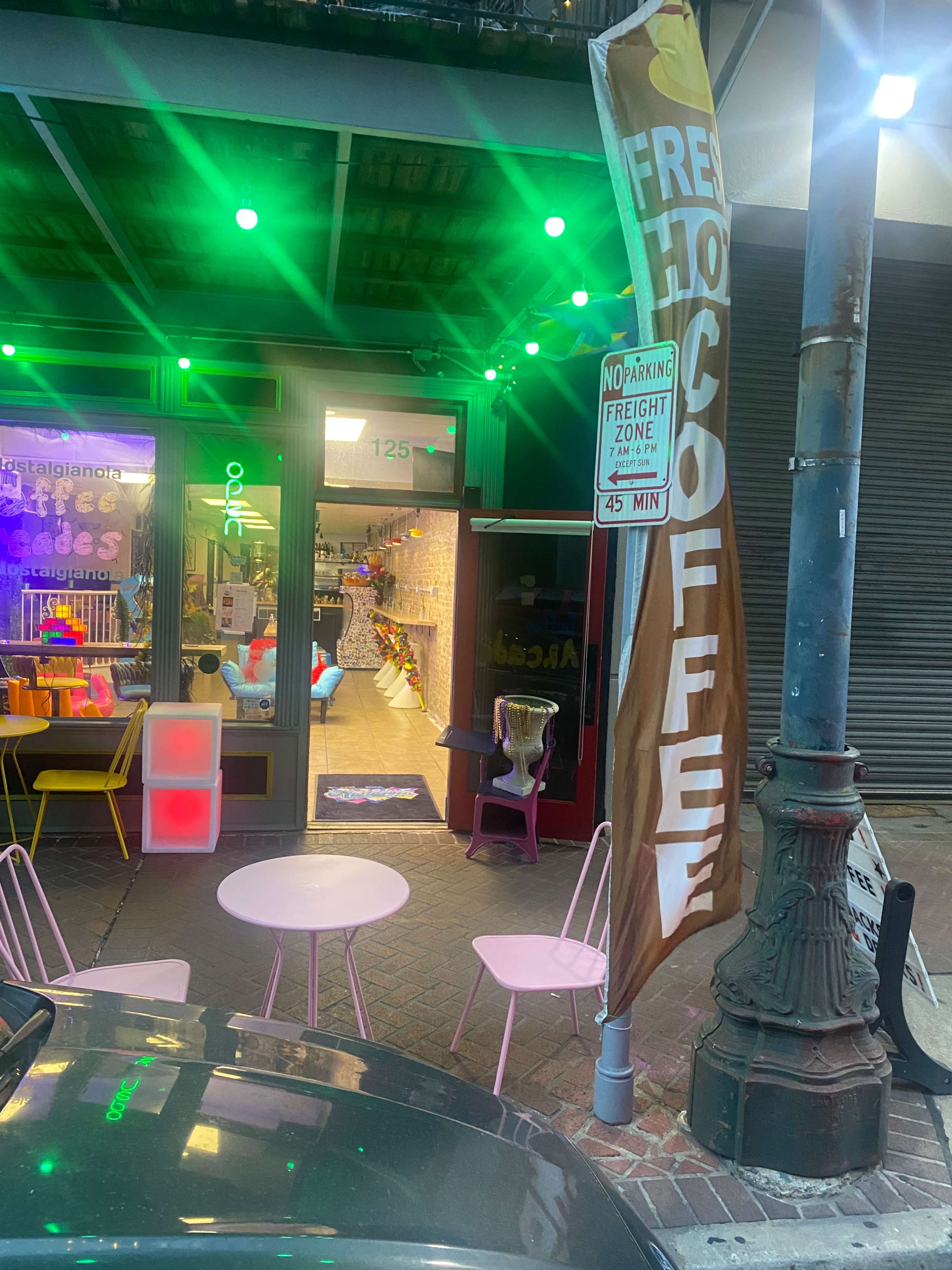 A café entrance with a large sign advertising "FREE HOT COFFEE," outdoor seating with pink chairs, and colorful lights illuminating the space.