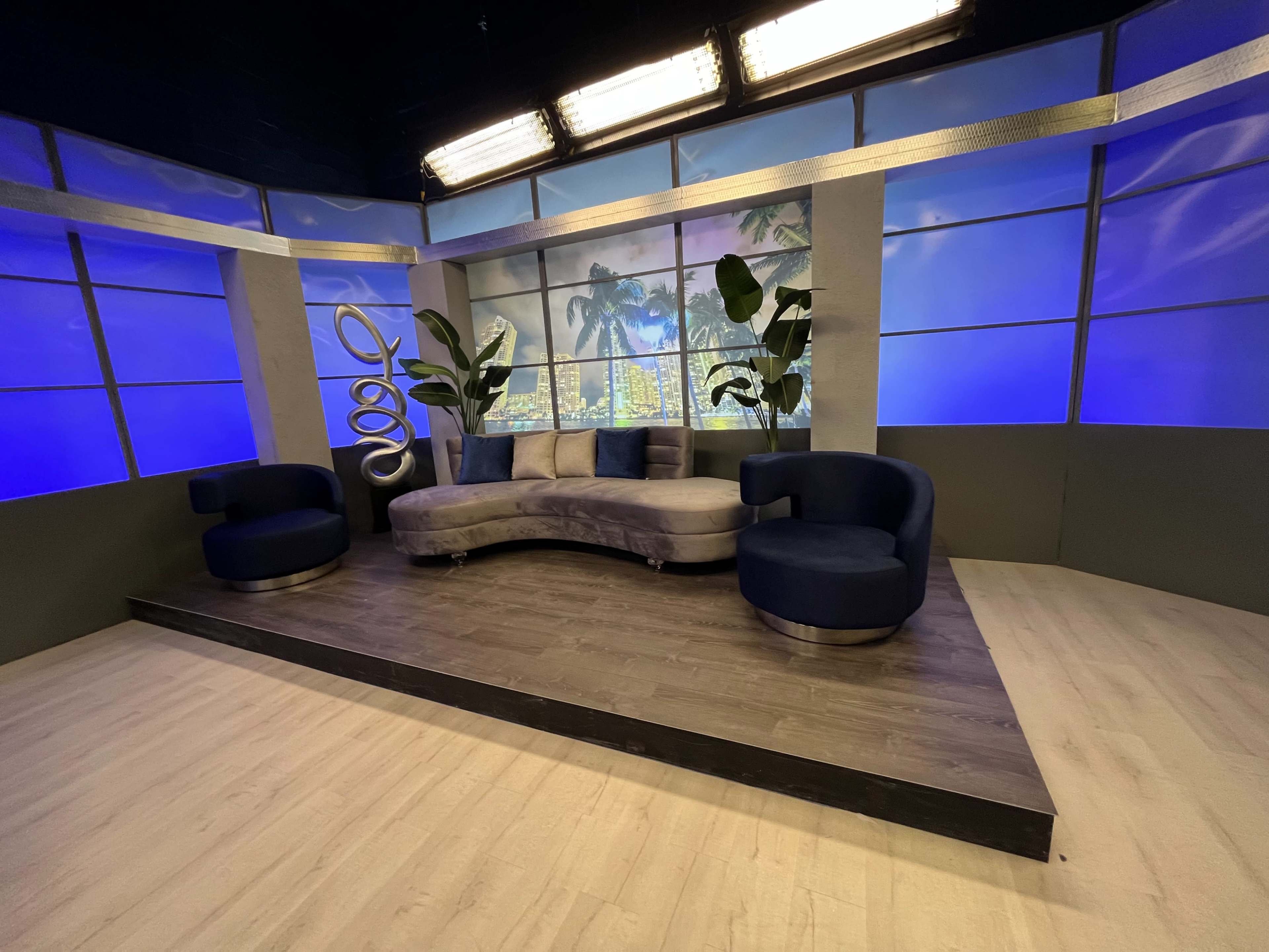 A modern television studio set featuring a curved sofa, two chairs, and large windows with a night cityscape backdrop.