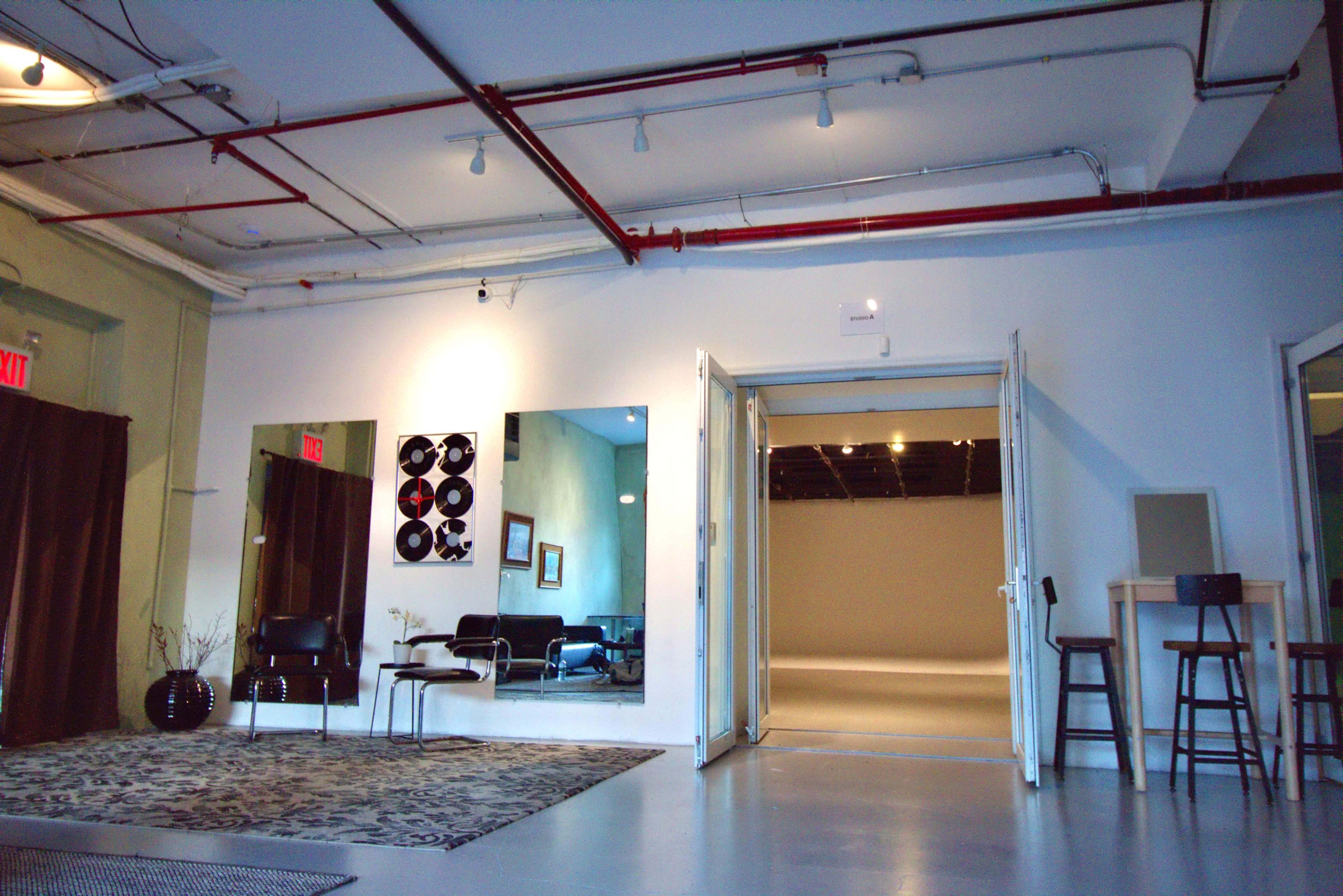The image shows a spacious interior with a wooden floor, featuring a seating area, mirrors on the walls, and an open doorway leading to a well-lit area.