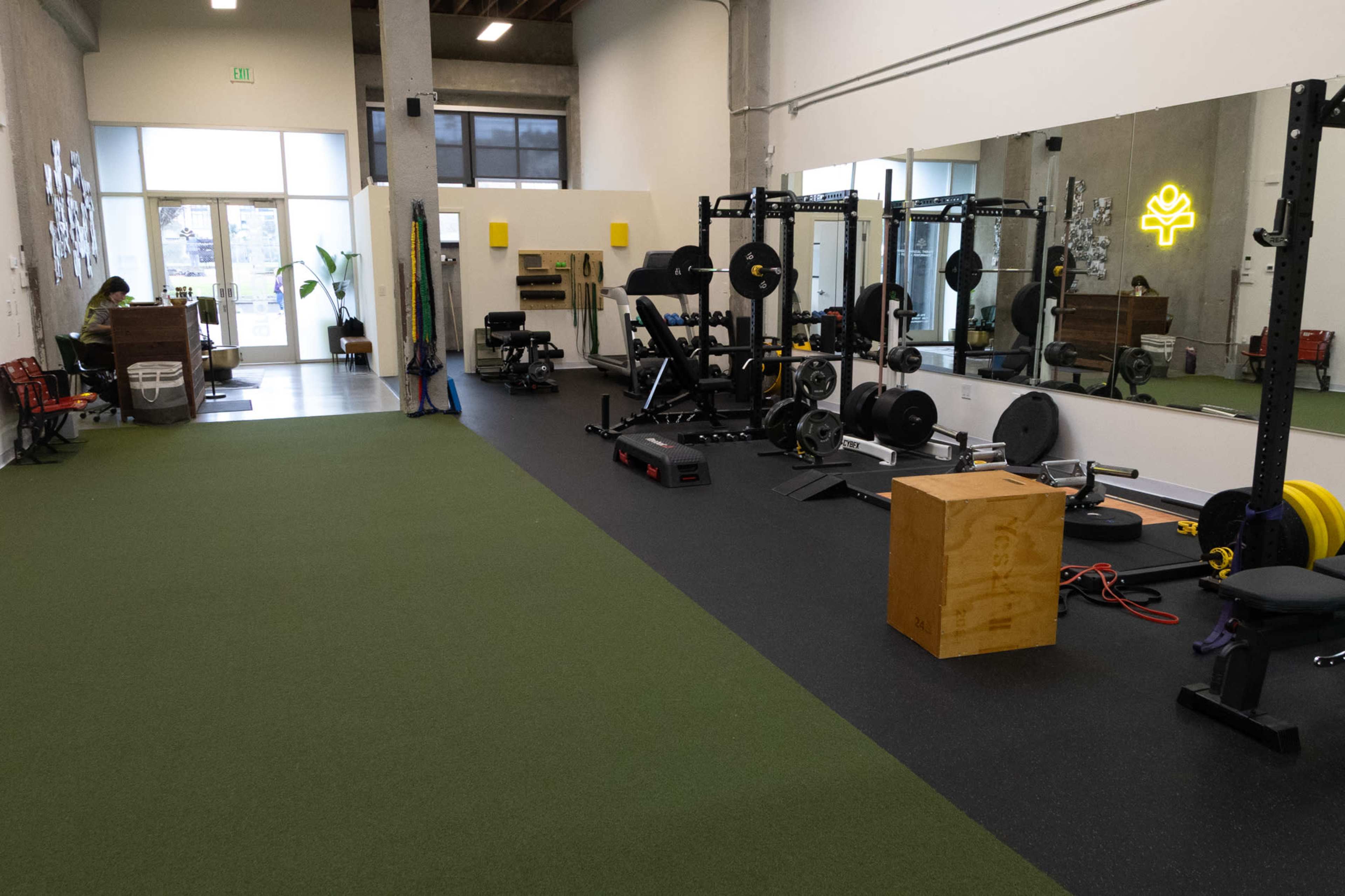 The image shows a gym interior featuring various weightlifting equipment, exercise mats, and a large mirror along one wall.