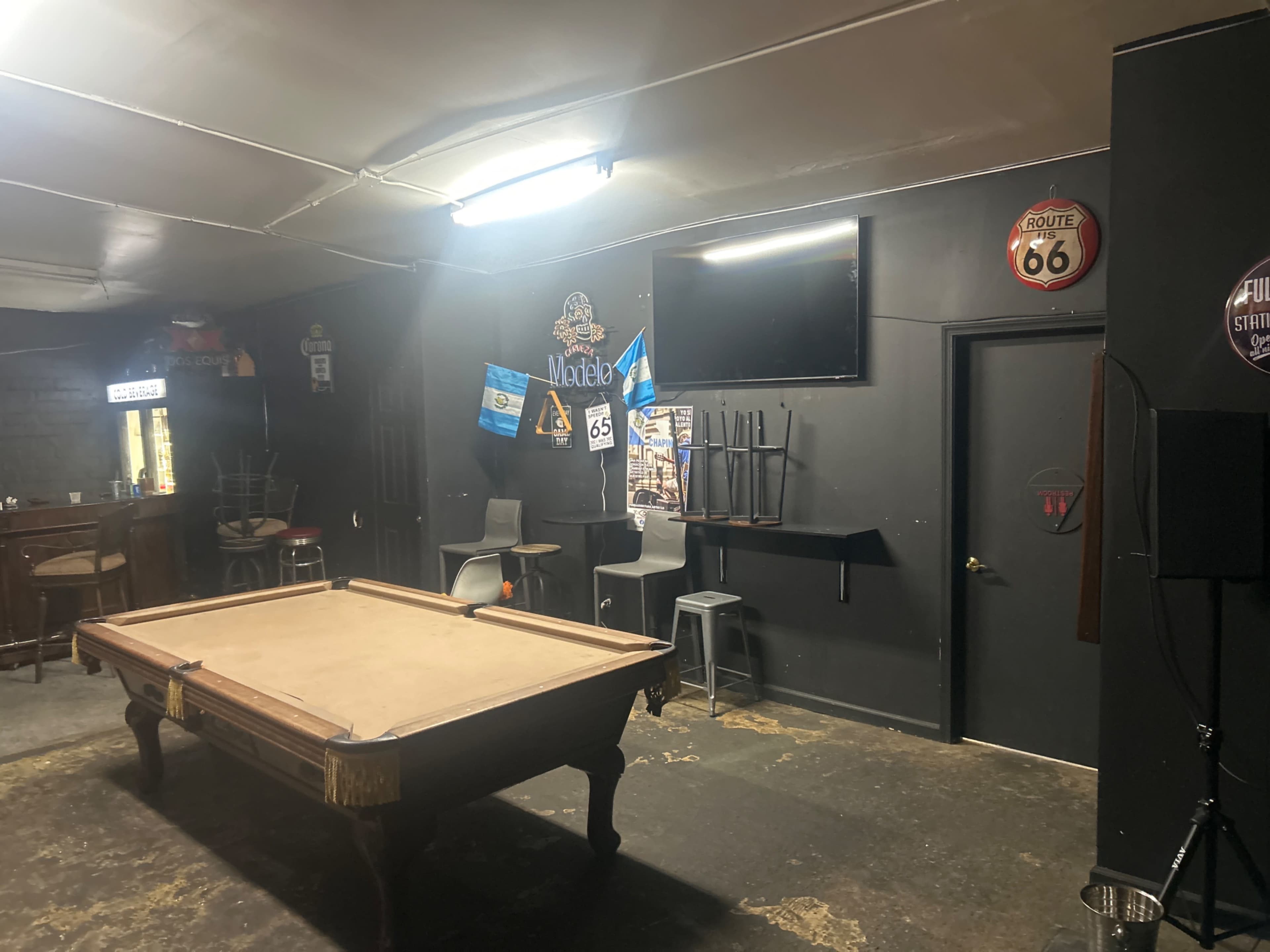 The image shows a dimly lit room featuring a pool table, bar stools, a television mounted on the wall, and various decorations including Route 66 memorabilia.