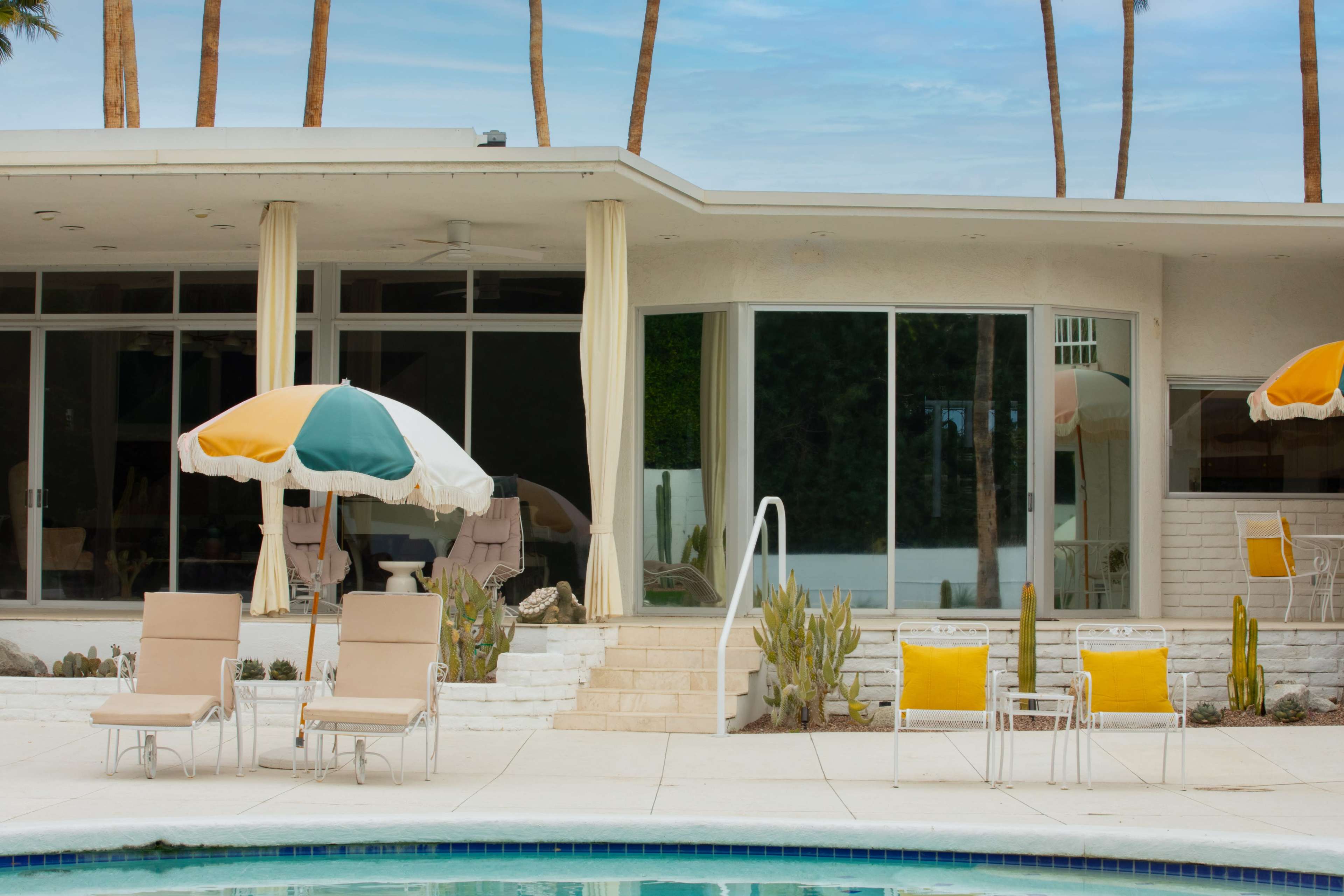 The image showcases a modern, mid-century home with large glass windows, a swimming pool in the foreground, and lounge chairs set up beside the water.