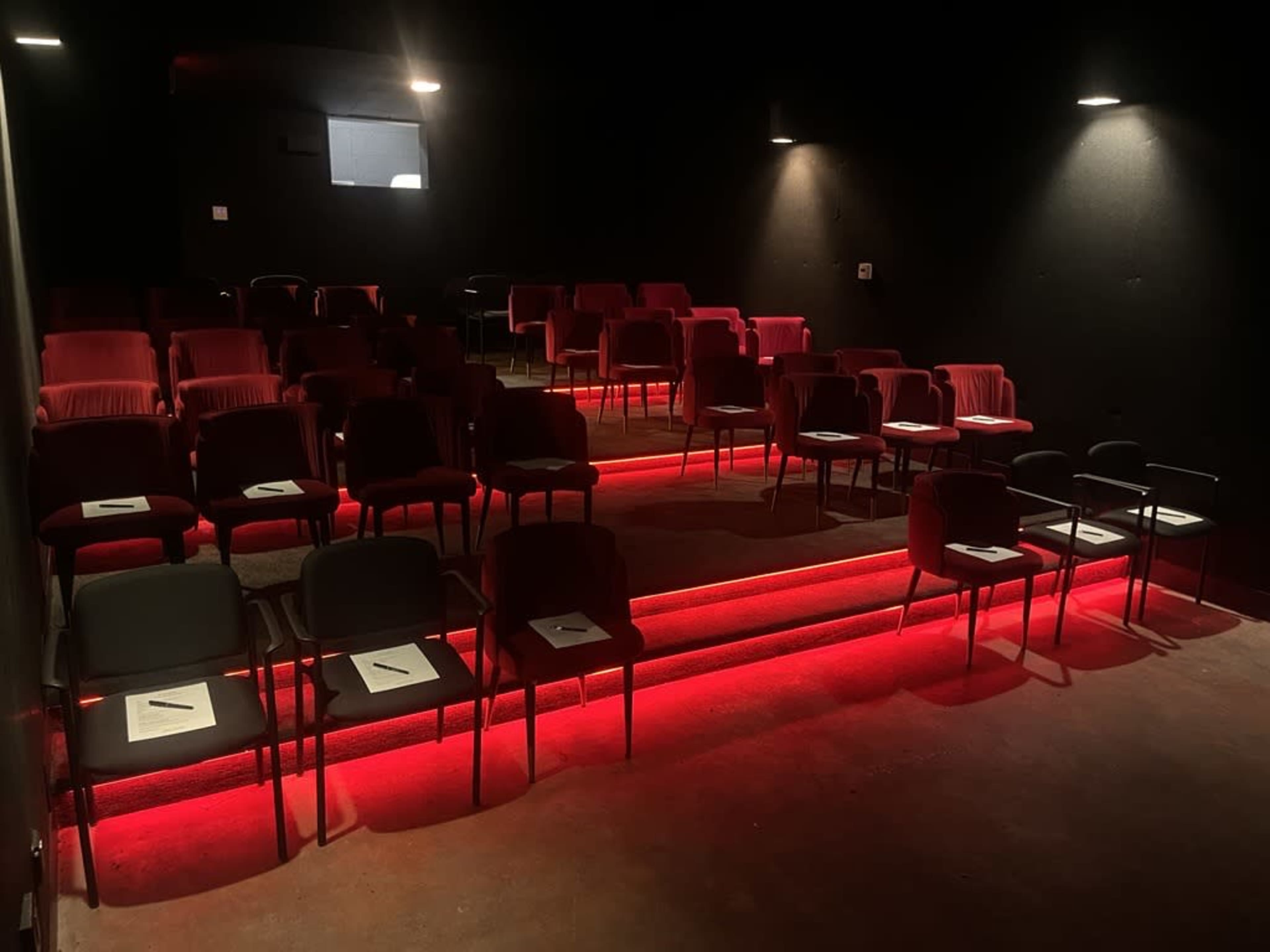 The image shows an empty theater space with red lighting illuminating the steps, and rows of chairs arranged with white papers on them.