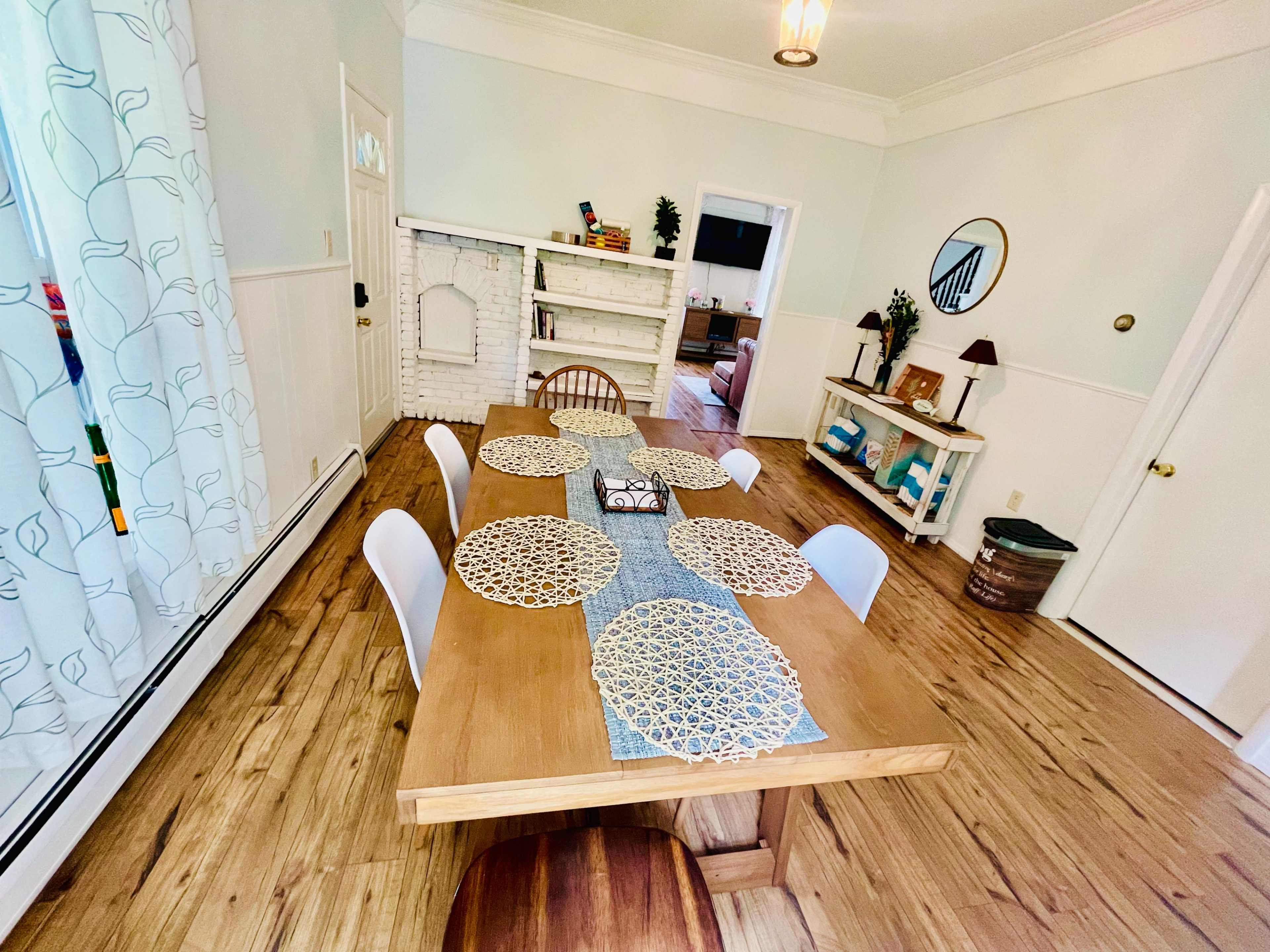 A wooden dining table is set with placemats, and a runner is placed centrally in a light-filled room featuring white walls and hardwood flooring.