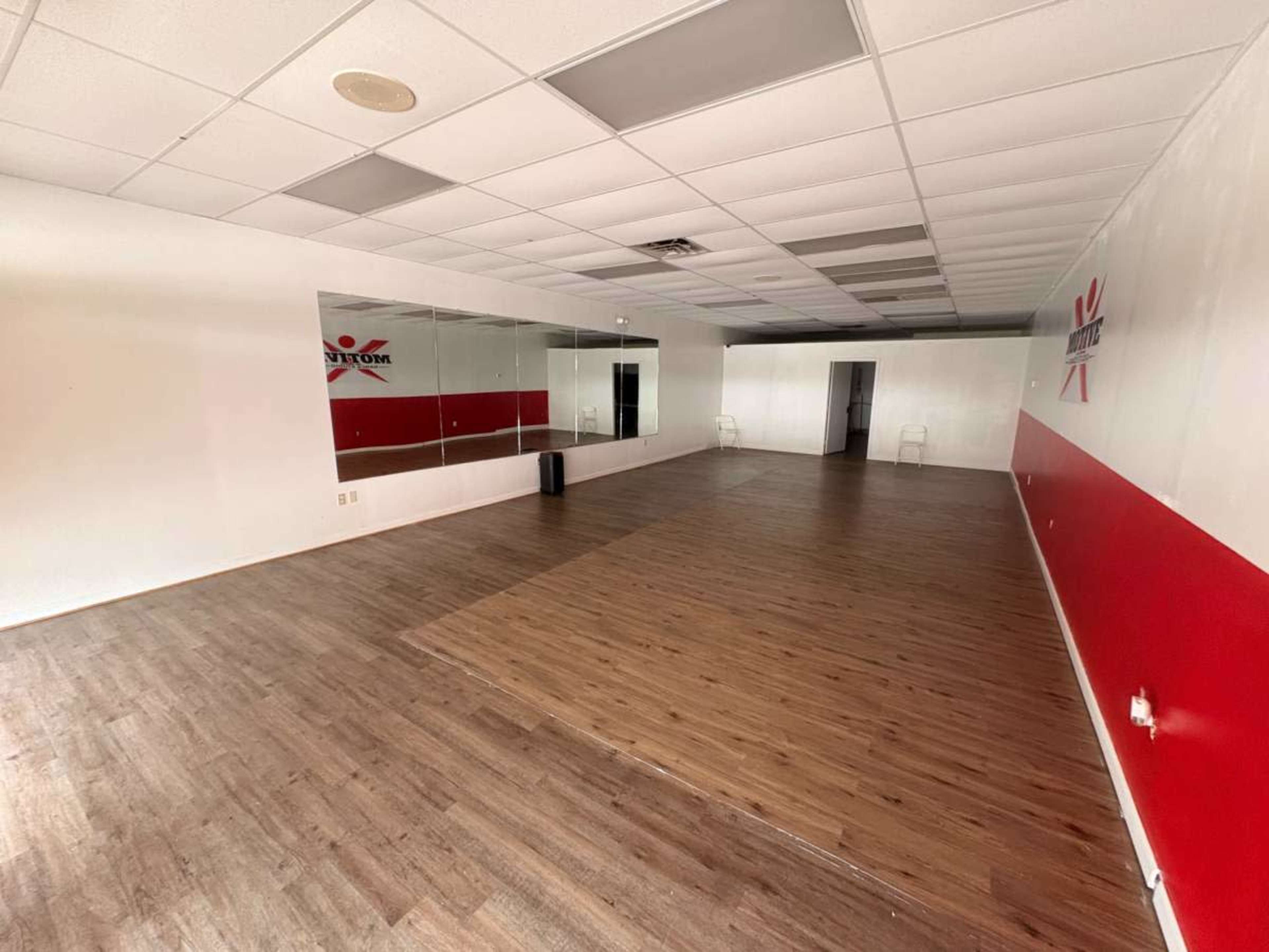 The image shows an empty room with wooden flooring, a red and white accent wall, mirrors on one side, and a couple of chairs against the wall.