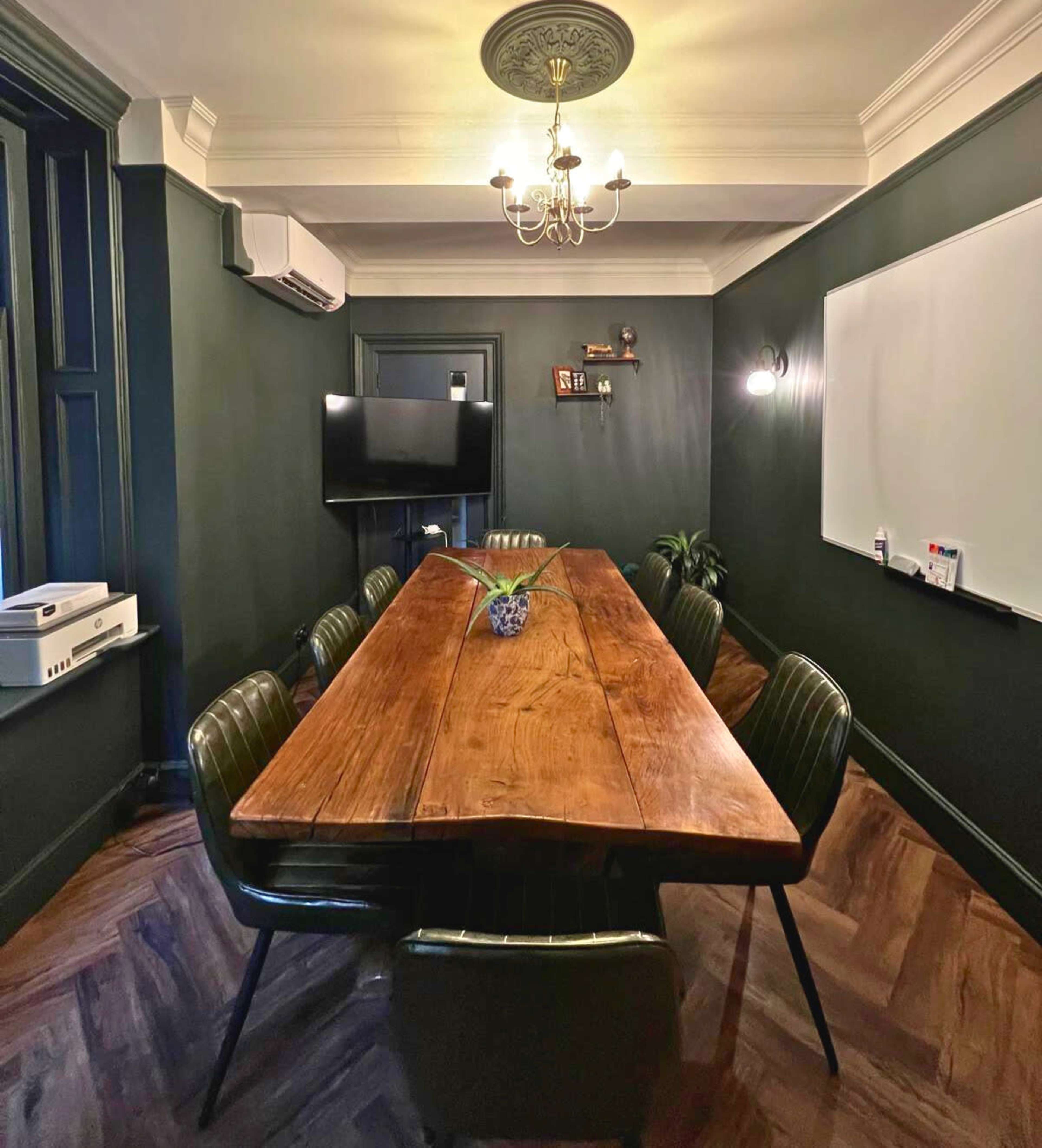 The image shows a conference room with a large wooden table, eight green upholstered chairs, a television mounted on the wall, and a whiteboard.
