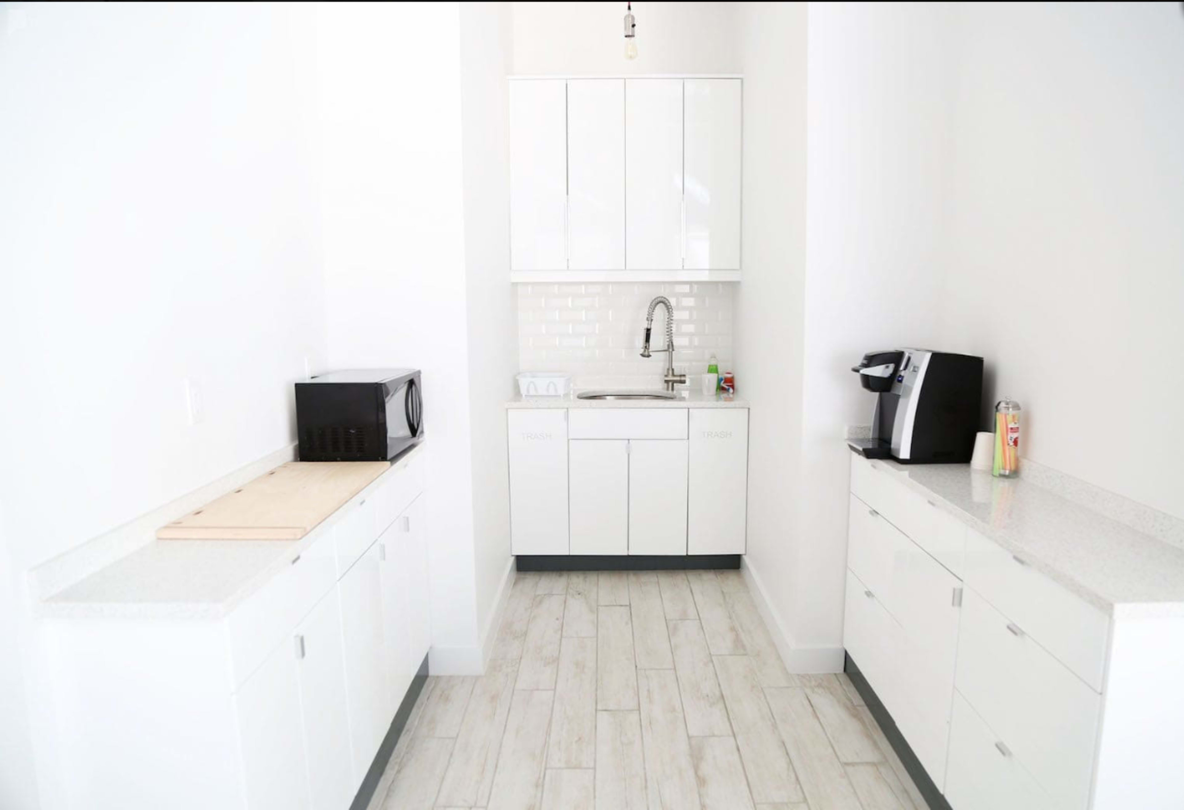 The image shows a modern kitchen space featuring white cabinetry, a sink, a microwave, and a coffee machine, with light-colored flooring.