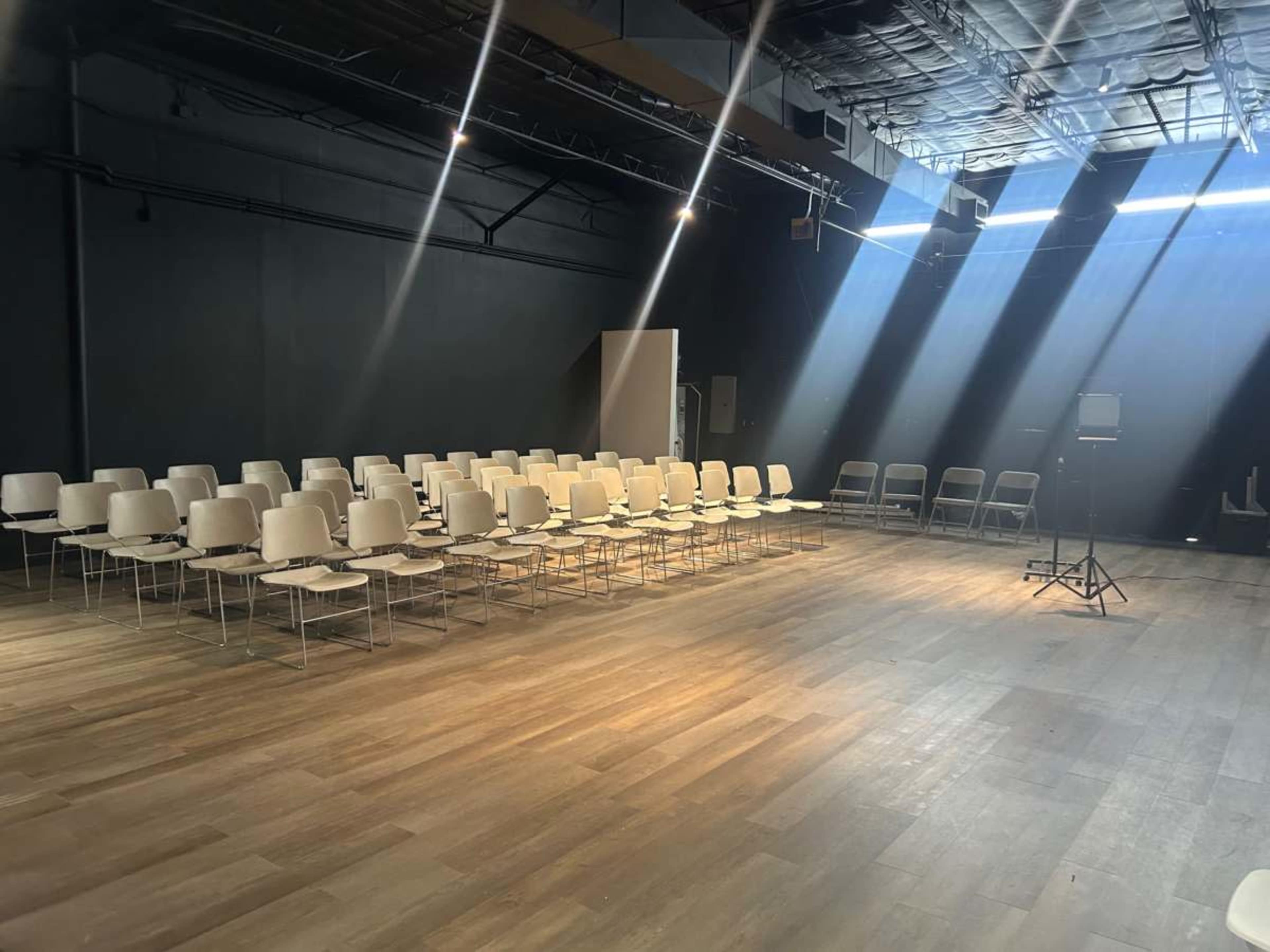 The image shows a spacious room with rows of white chairs arranged for an event, illuminated by beams of sunlight coming through the ceiling.
