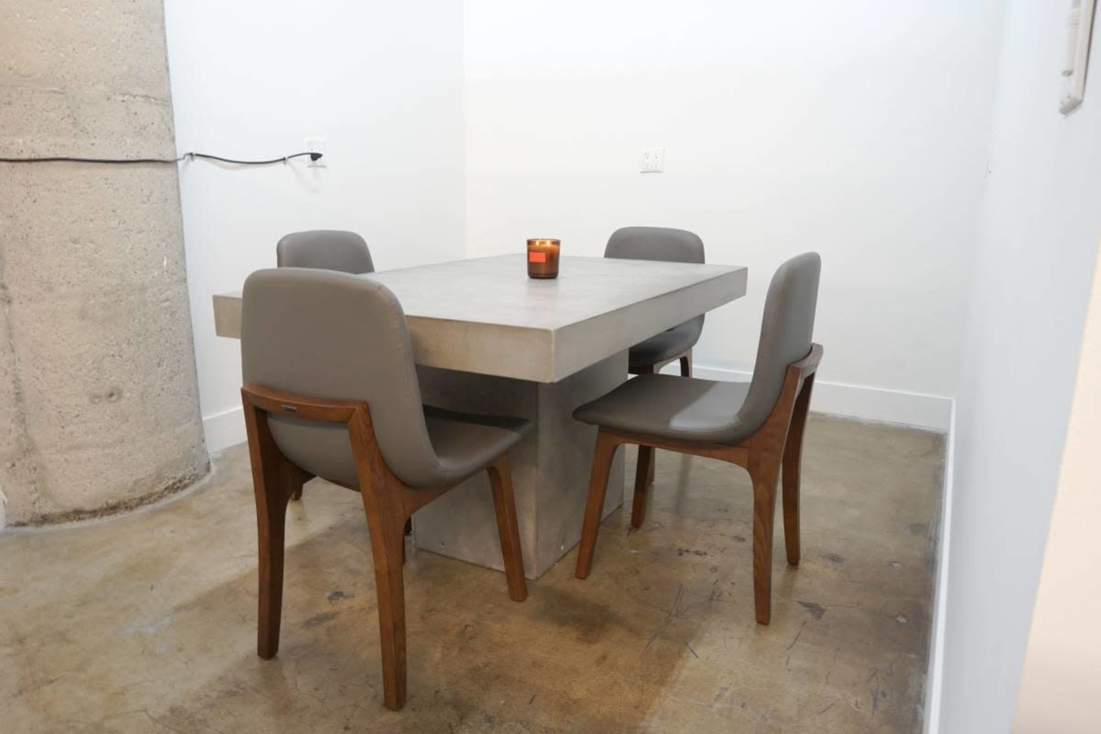 A minimalist dining table with a concrete top and wooden legs is surrounded by four upholstered chairs in a simple, empty room.