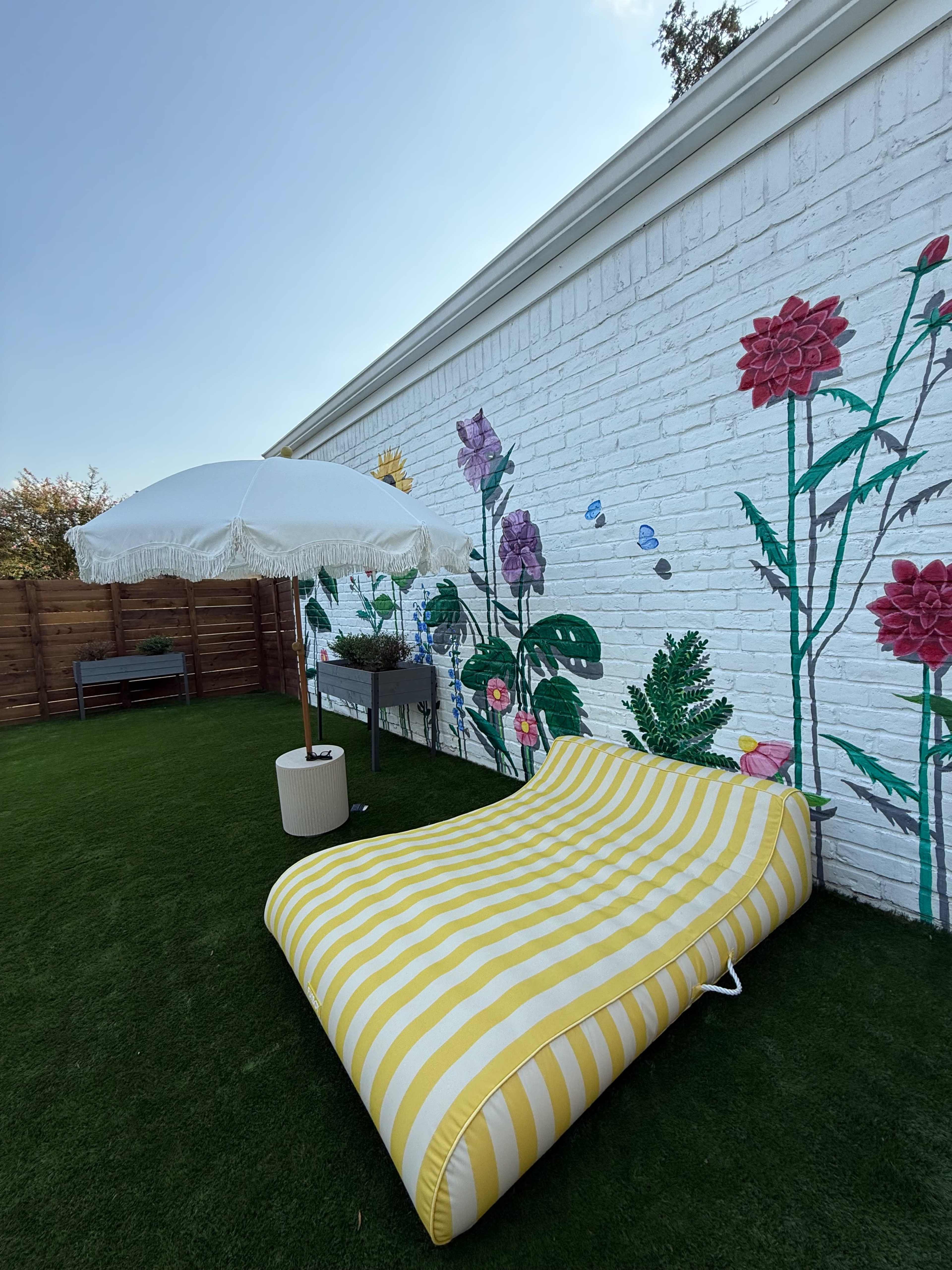 A large yellow-striped lounge chair sits beside a white umbrella in front of a brick wall decorated with colorful floral murals.