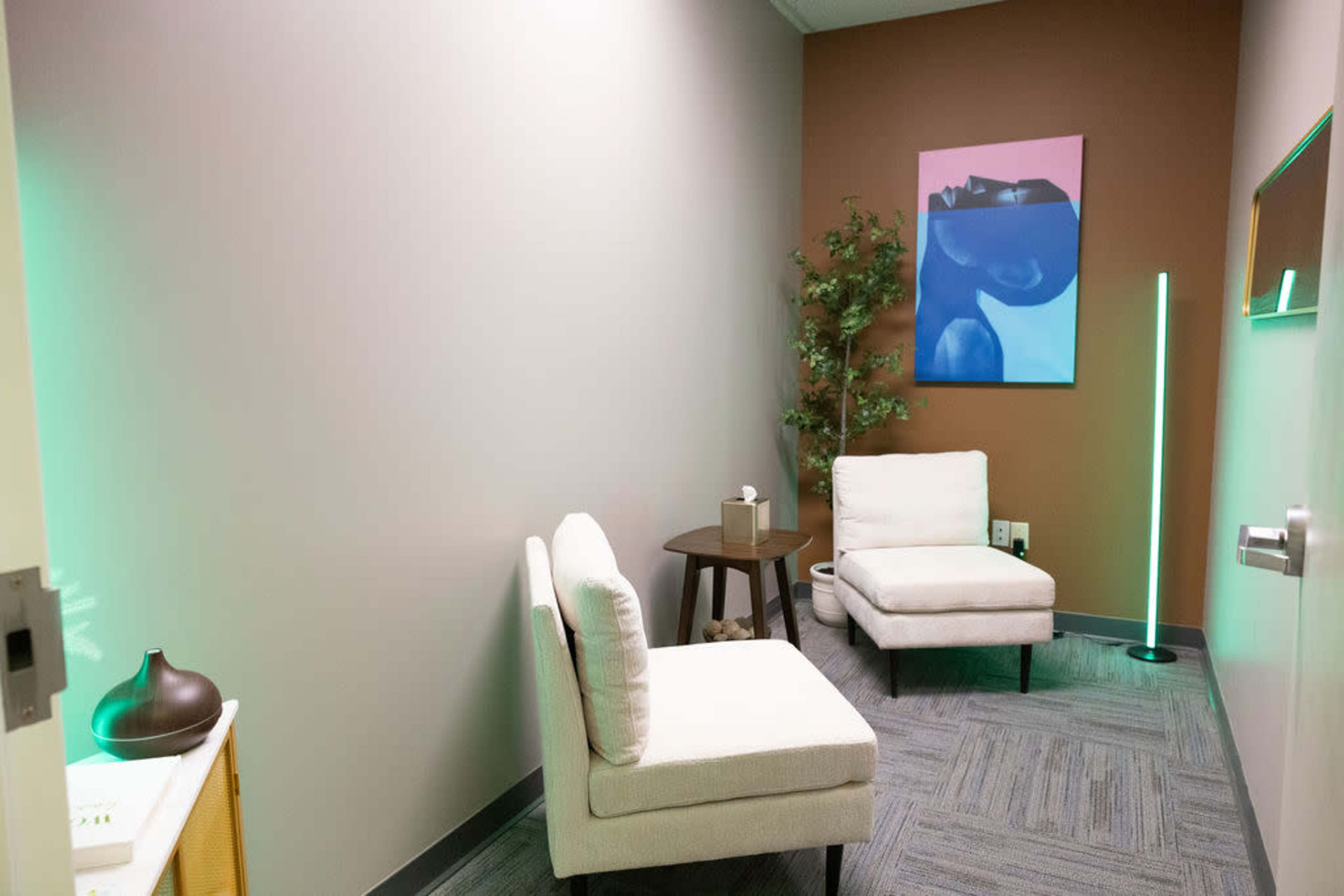 The image shows a small, modern therapy room with two chairs, a table, a decorative plant, and a colorful artwork on the wall.