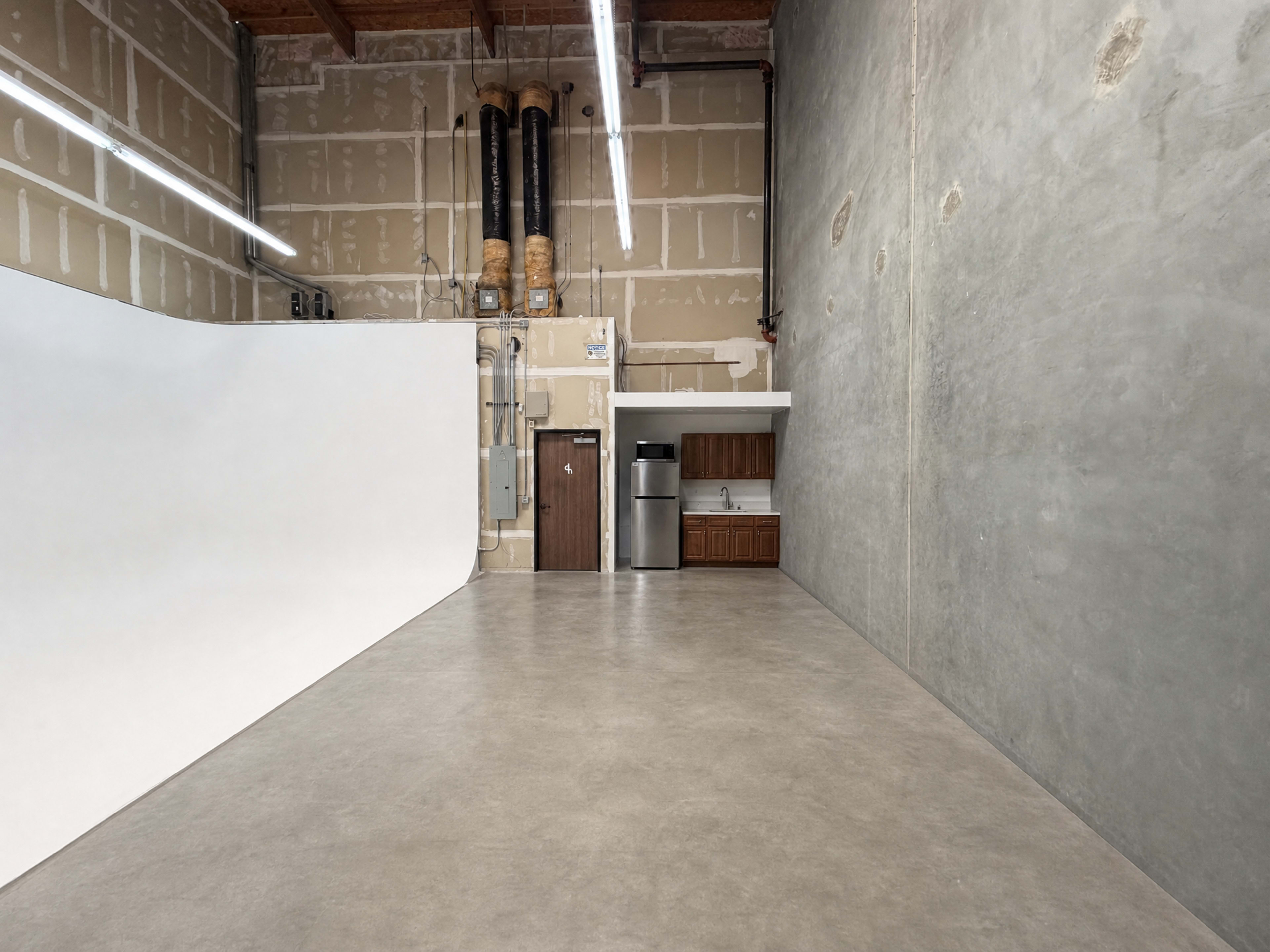 The image shows an empty industrial space with a white backdrop, a kitchenette area, and exposed pipes along the walls.