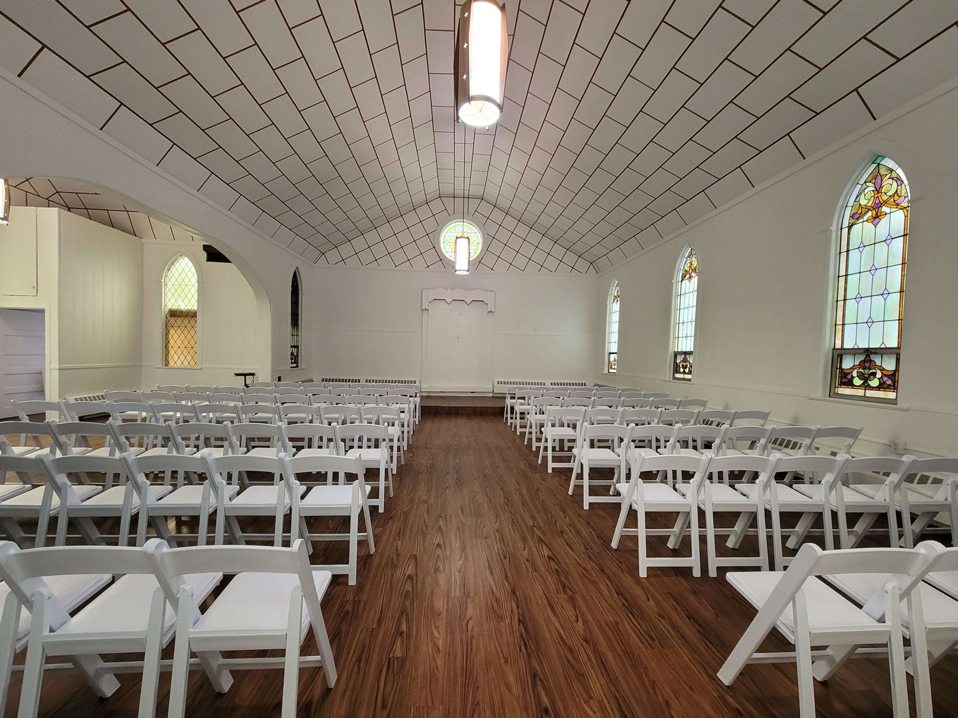 The interior of a chapel with rows of white chairs facing a recessed stage and stained glass windows along the walls.