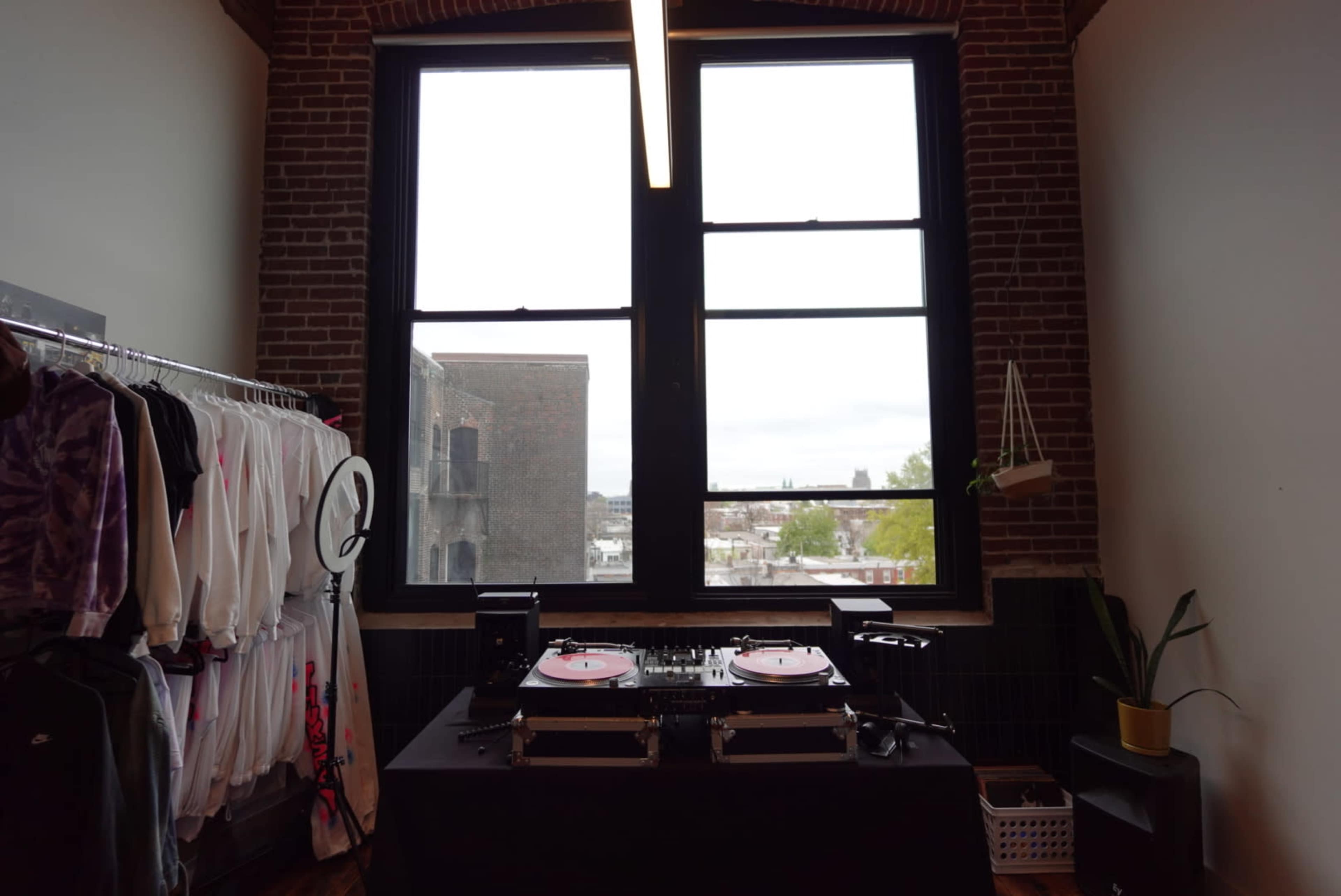 The image shows a room with large windows, a DJ setup with turntables on a black table, and a clothing rack filled with various garments.