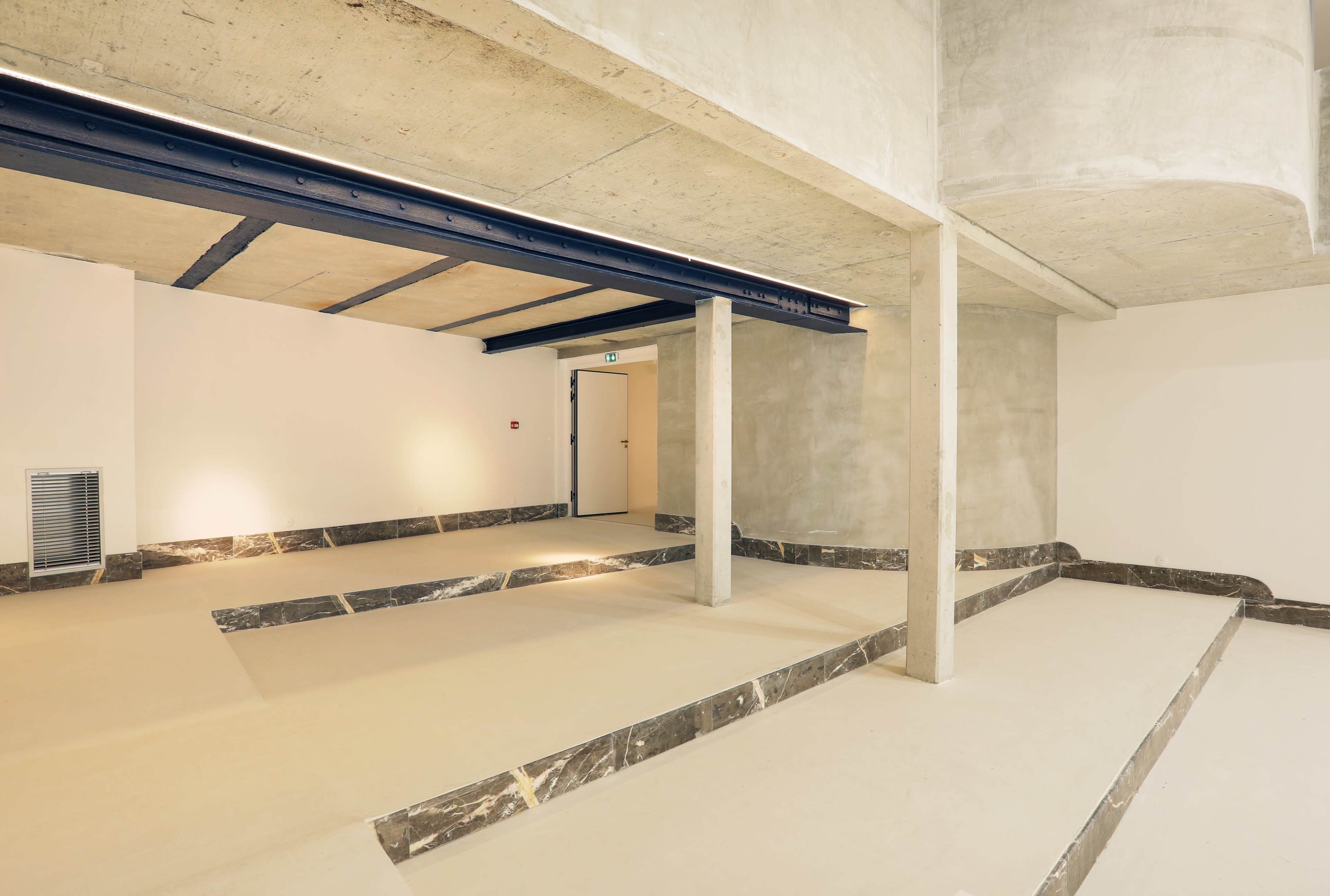 An interior space with concrete walls, exposed beams, and a floor featuring sunken areas lined with dark marble.