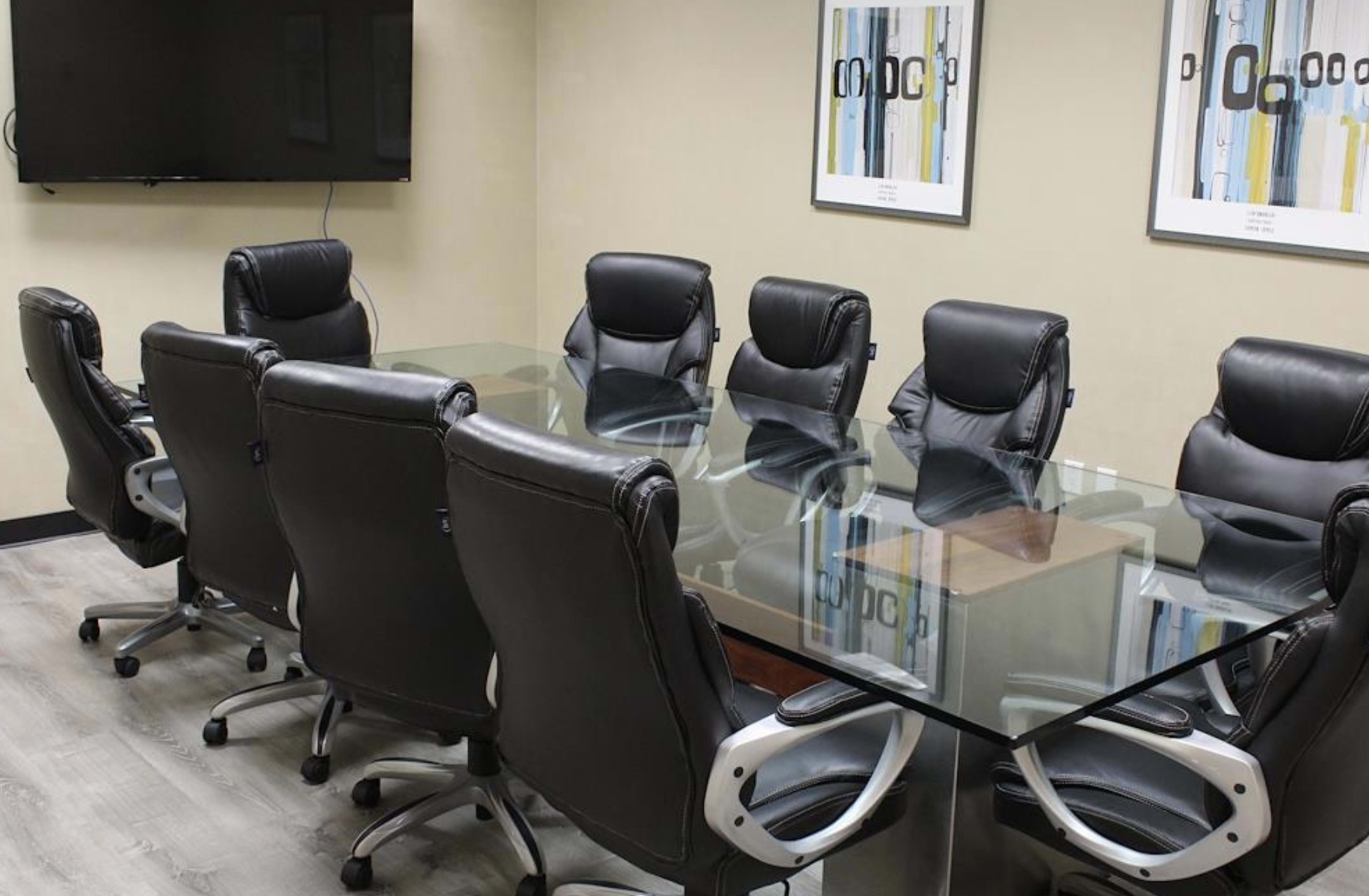 A conference room features a glass table surrounded by eight black ergonomic chairs, with a television mounted on the wall and framed art displayed.